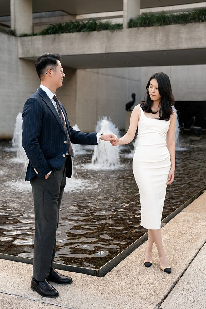 Dave look at Kath when hands holding in front of a fountain