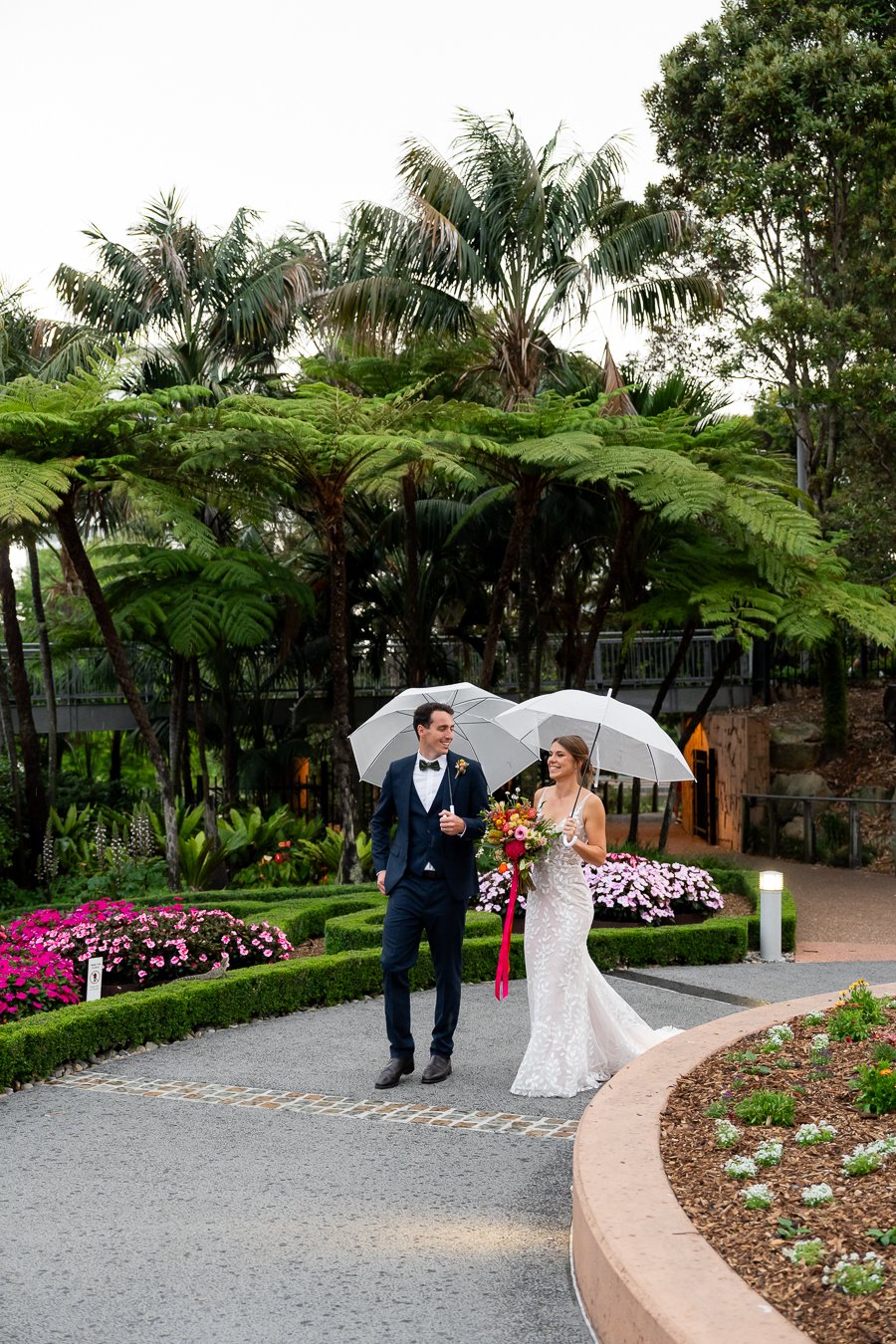 bride and groom hold umbrella walking in the garden with laugh