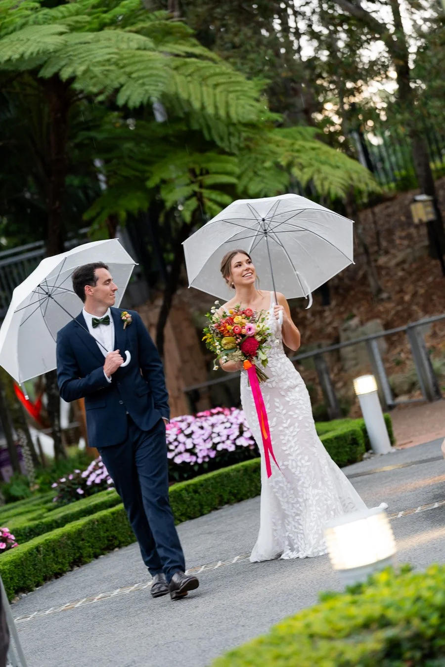bride and groom holding umbrellas walking inside a garden