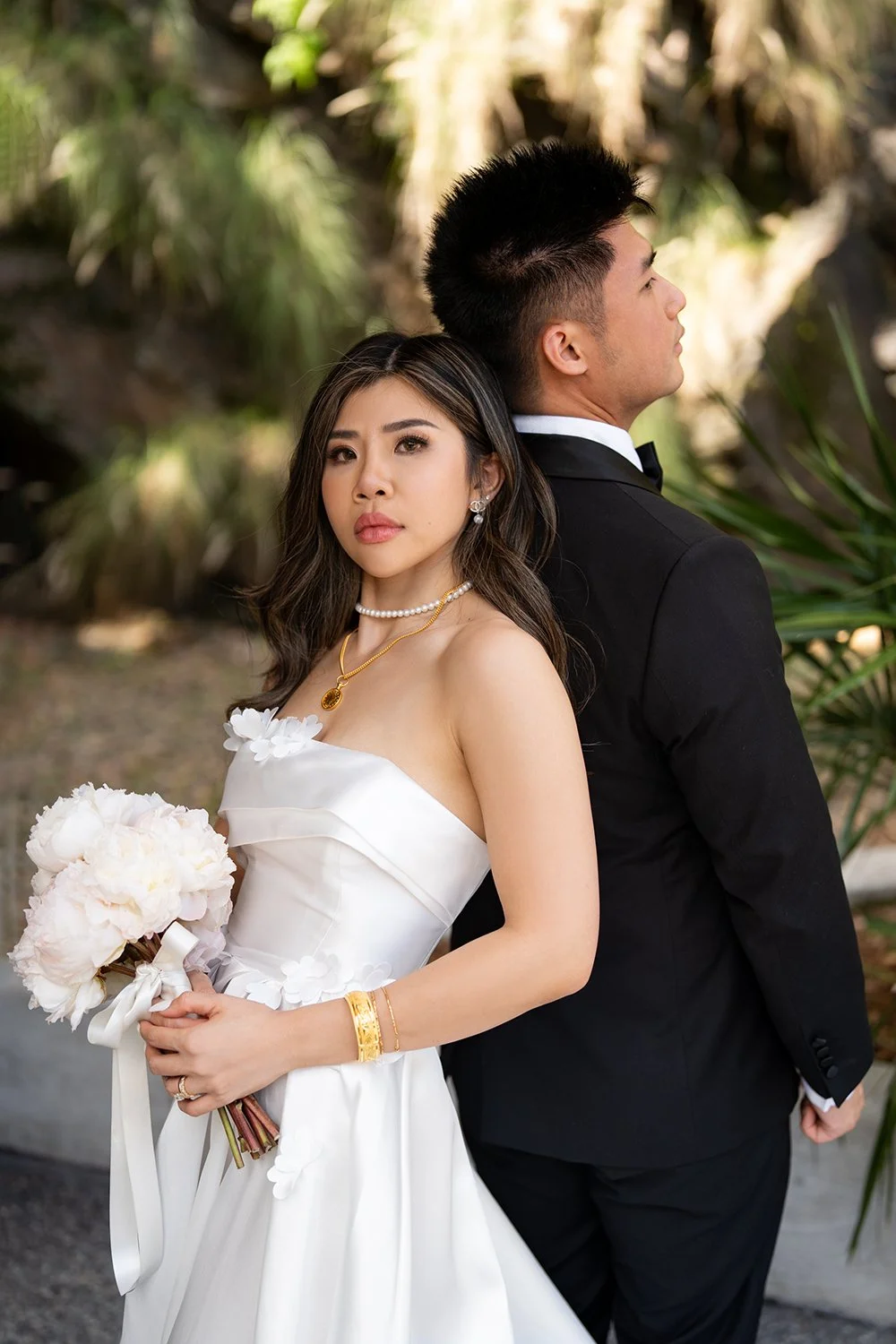 Bride Tiffy leaning back against the groom kai, looking at the Brisbane wedding photographer while holding her bouquet with both hands. The groom supports her gently, creating a sense of stability and trust. The backdrop of rugged rock stones enhance