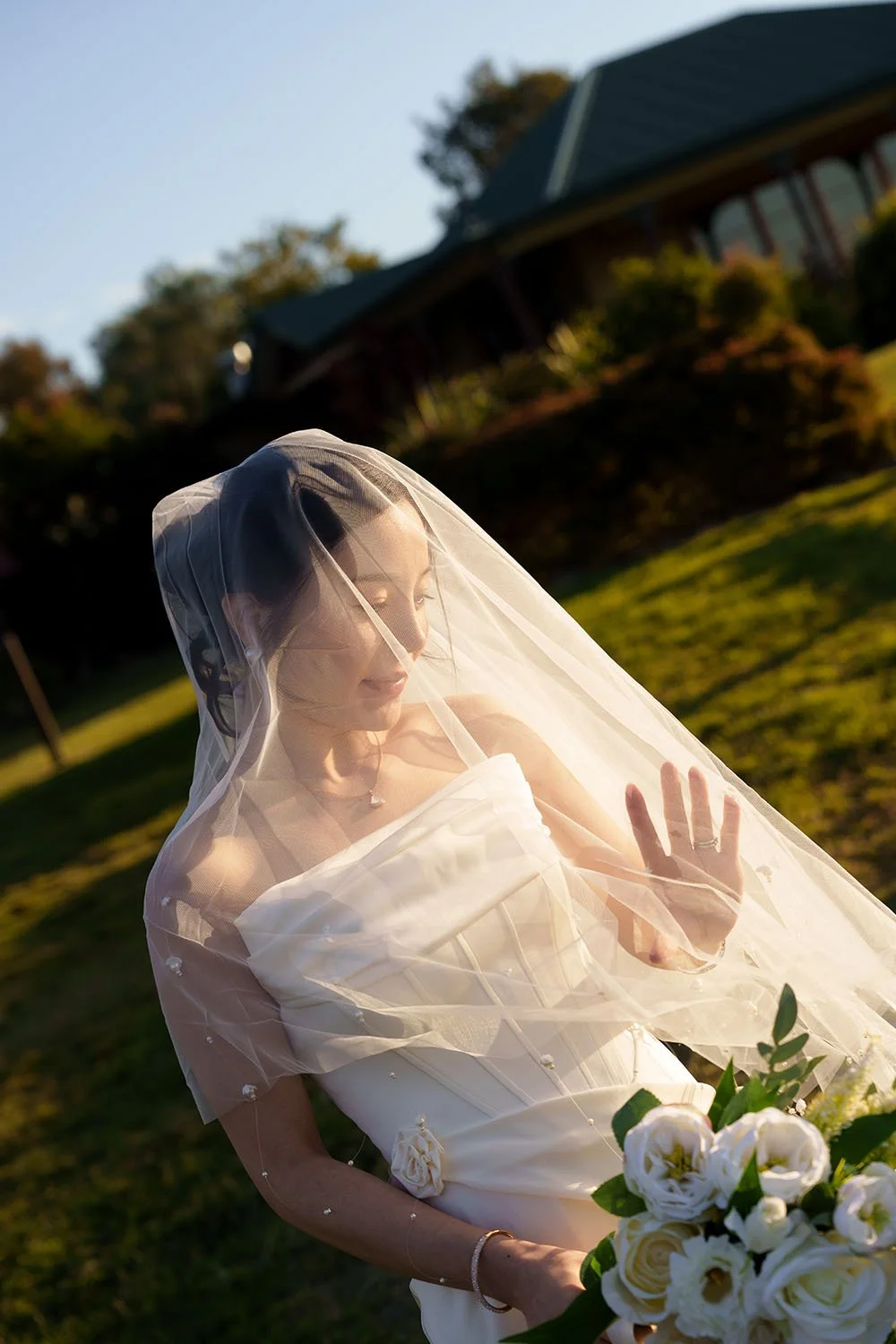 With Flora veil covering her head, the bride gently supports it with her hand, capturing a quiet and delicate moment of anticipation.