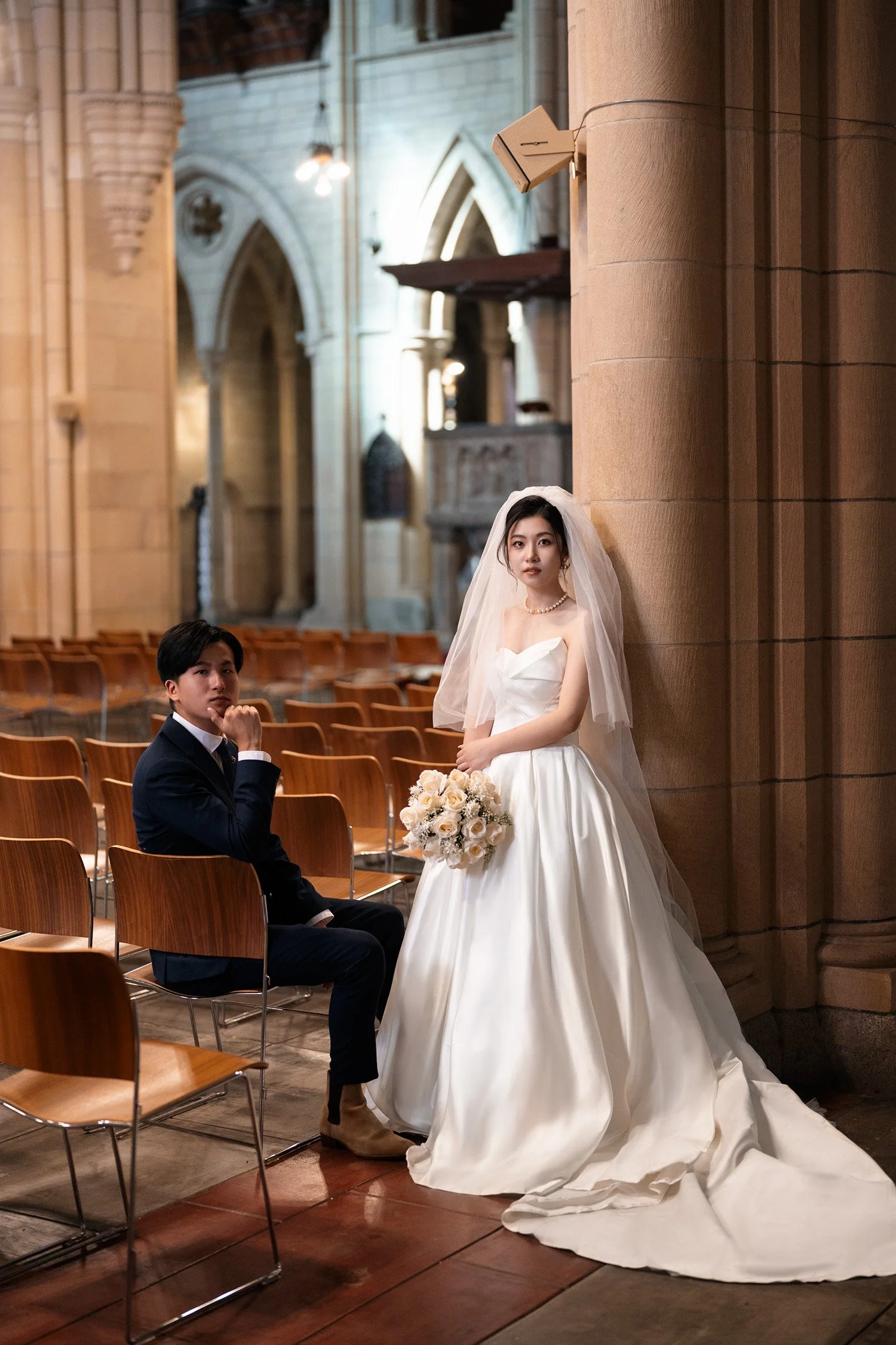 bride Cassie leaning on the column and groom Leo sitting on the chair both look at wedding photographer