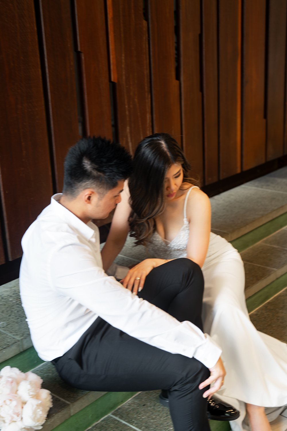 Tiffy and Kai sitting next to each other, with the bride gently holding the groom's leg as they both look down, creating a peaceful and quiet moment. The bride is in her elegant wedding gown, and the groom is dressed in a classic suit, exuding a sens