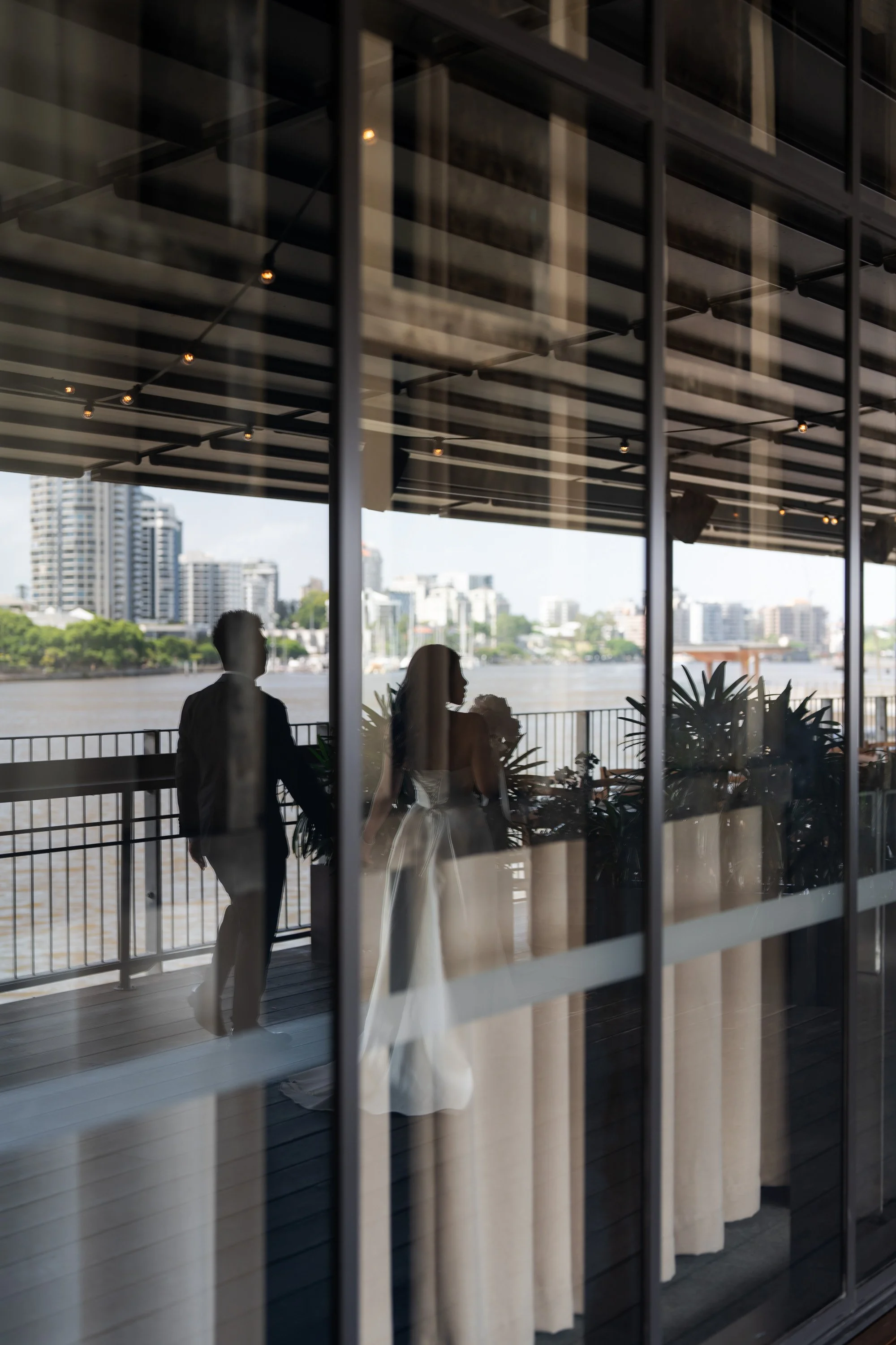 Bride Tiffy and groom Kai holding hands and walking together, captured through a mirror reflection. The reflection reveals the surrounding building and Brisbane River, adding depth to the romantic moment. The couple's love and connection are beautifu