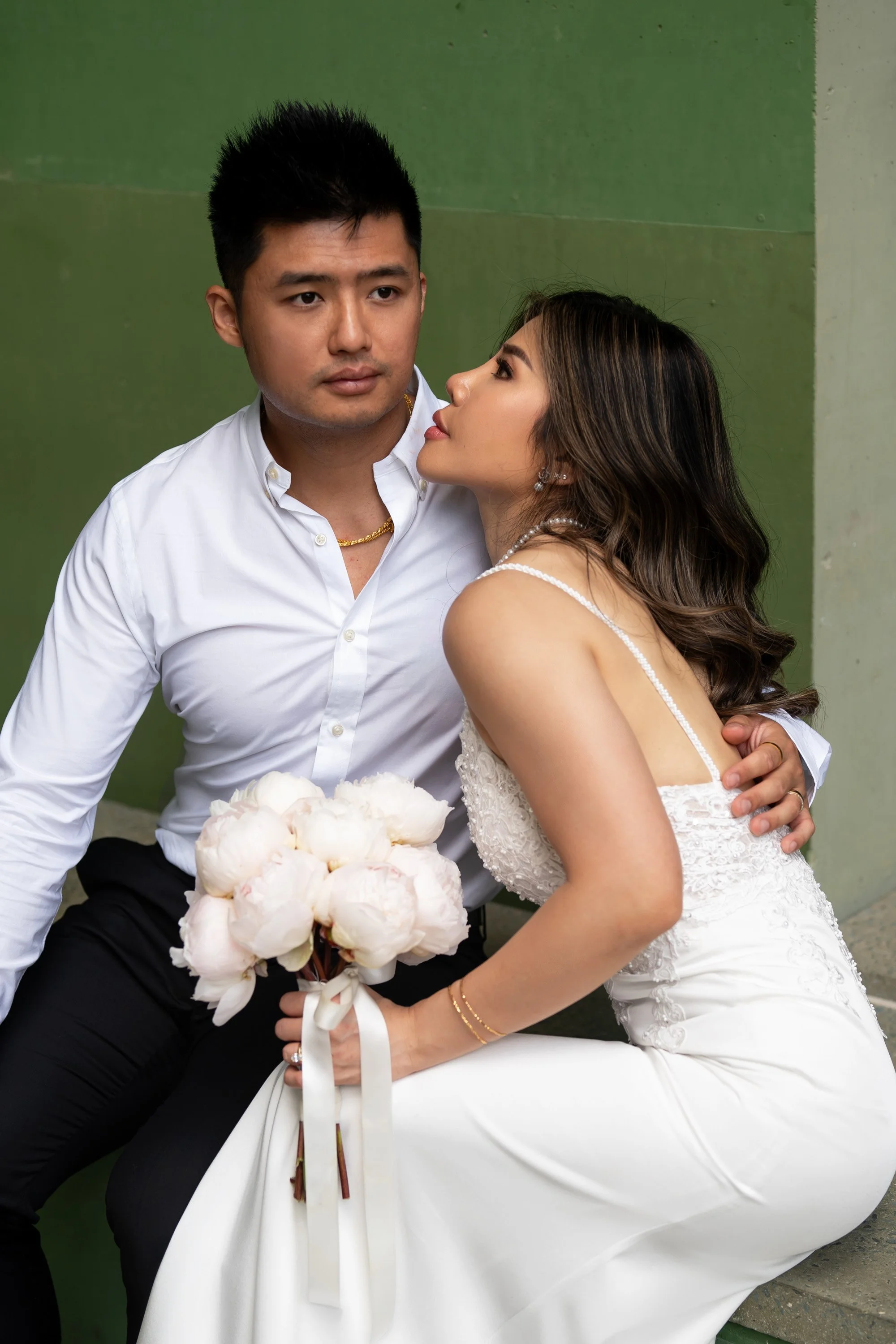 Groom Kai holding the bride Tiffy as they sit together, with the bride gazing lovingly at him. The bride is dressed in an elegant wedding gown, and the groom in a classic suit, creating a tender and intimate moment that highlights their deep connecti