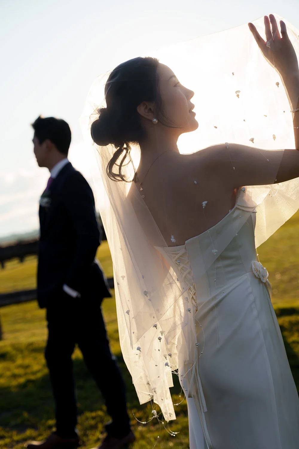 veil covering Flora's head, and her hands support the veil inside, and look it. groom Eric hands inside the pants pockets 5 steps away from her