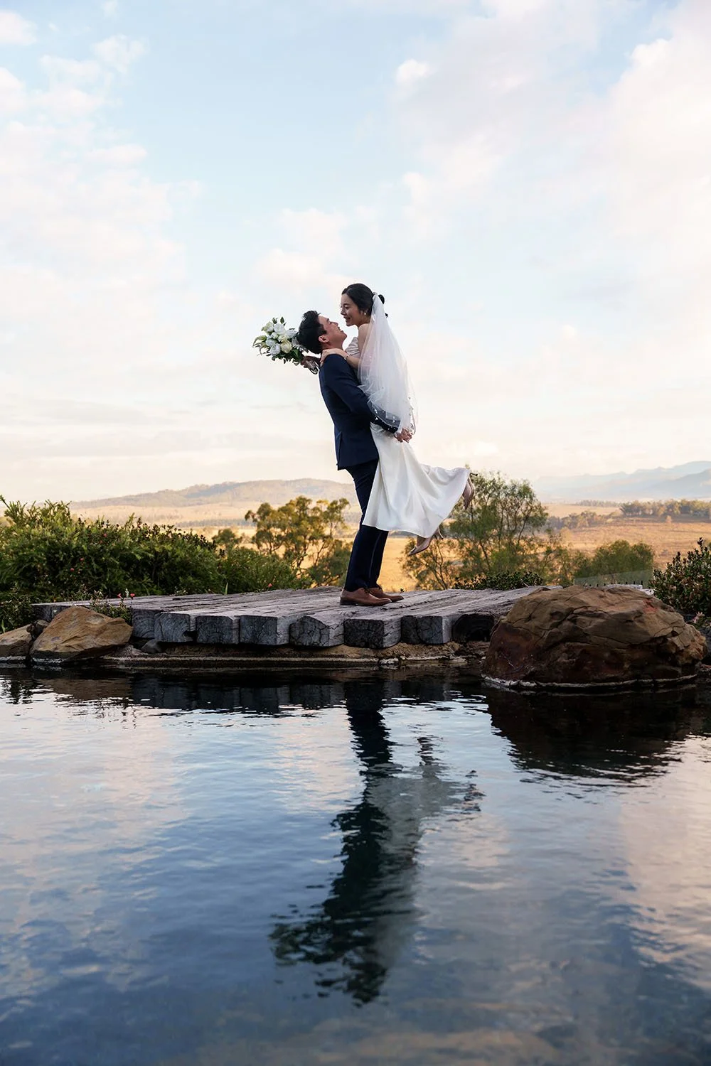 Eric holding up Flora and look at each other, while Flora banning her feet, both stand on a wooden stage next a water pool