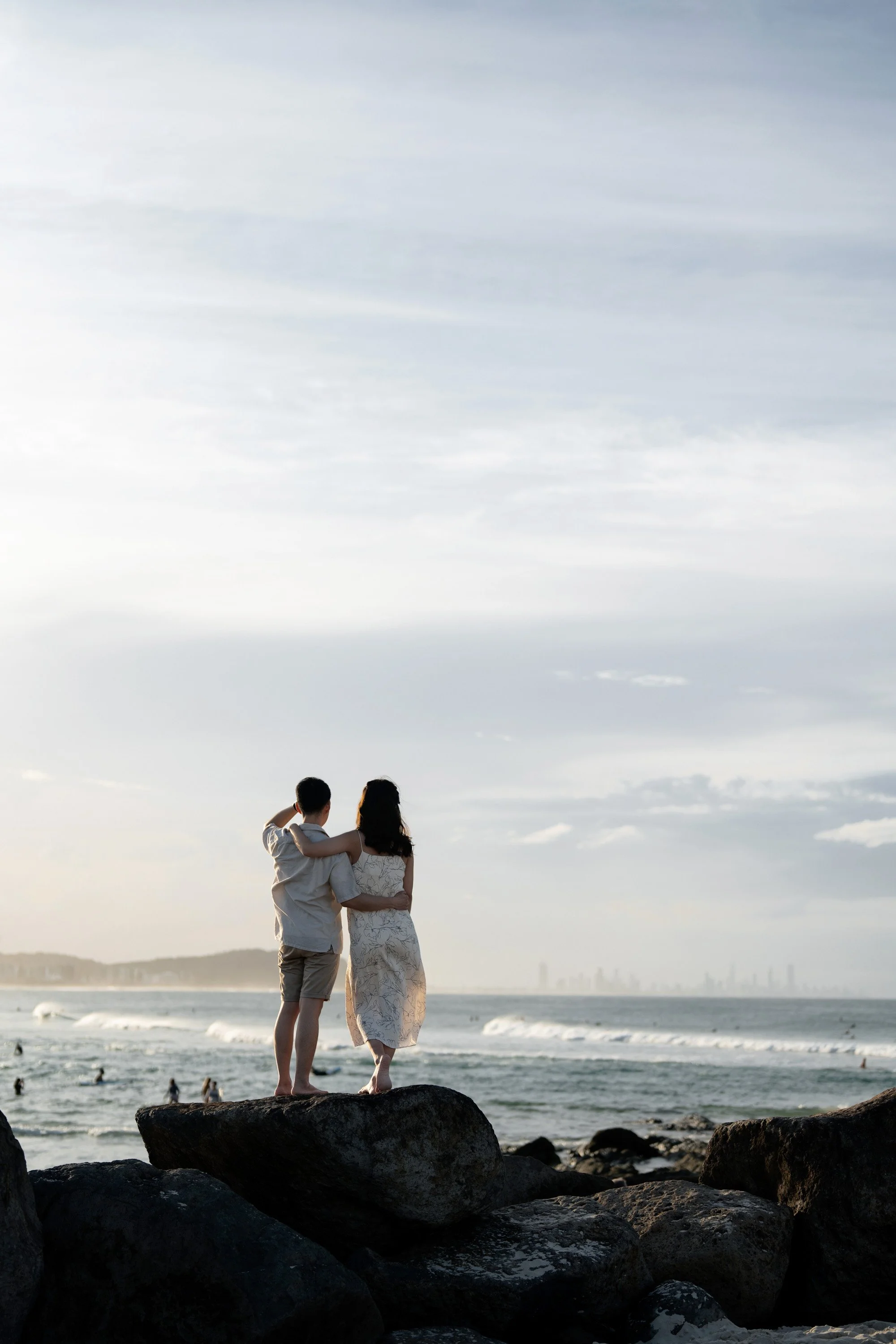 Paul and Krystel holding each other stand on the rock watch the Gold Coast view and people surfing