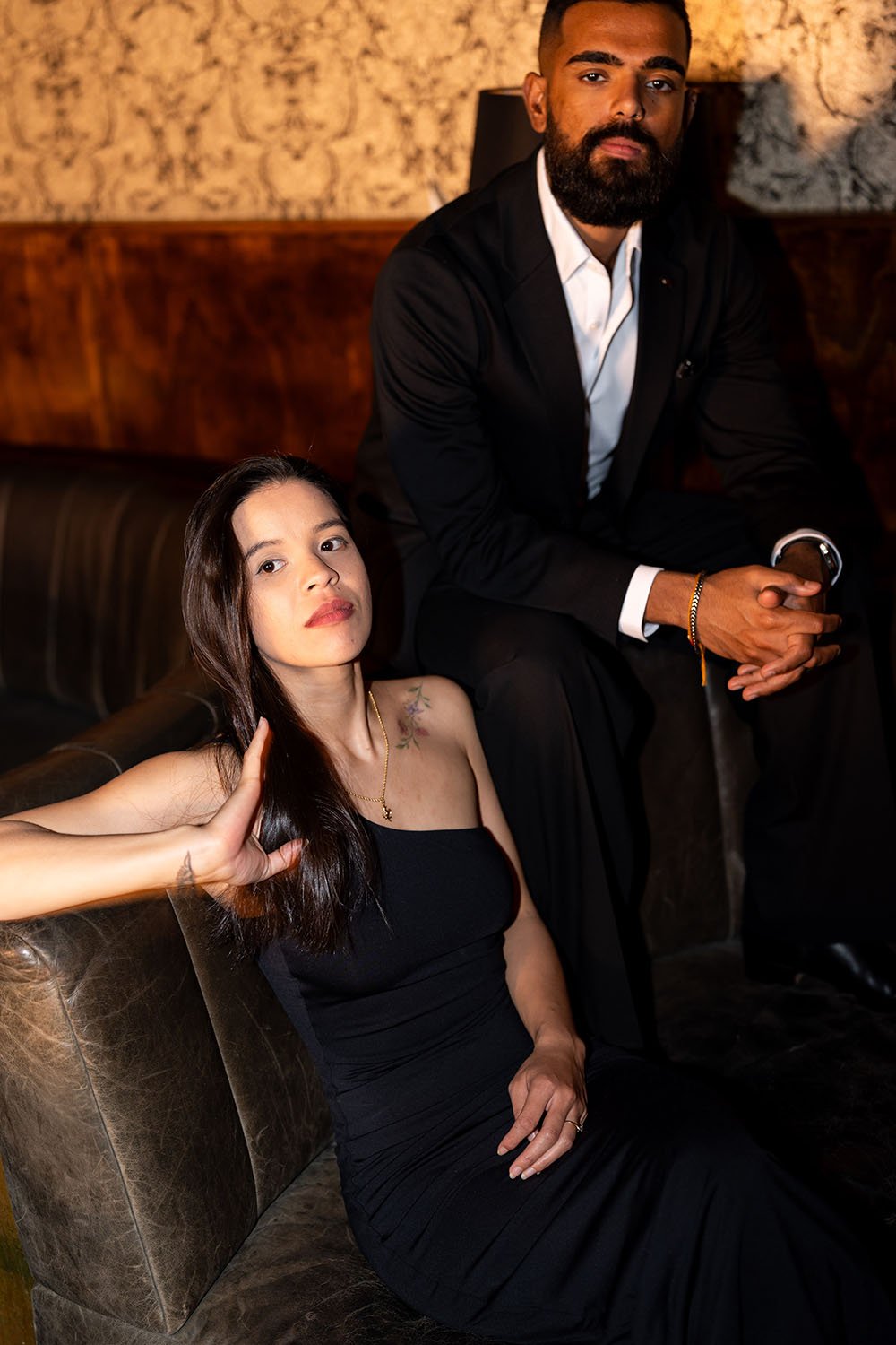 Jasmin sitting on a sofa playing with her hair and Jay sitting on the sofa arm look at the camera