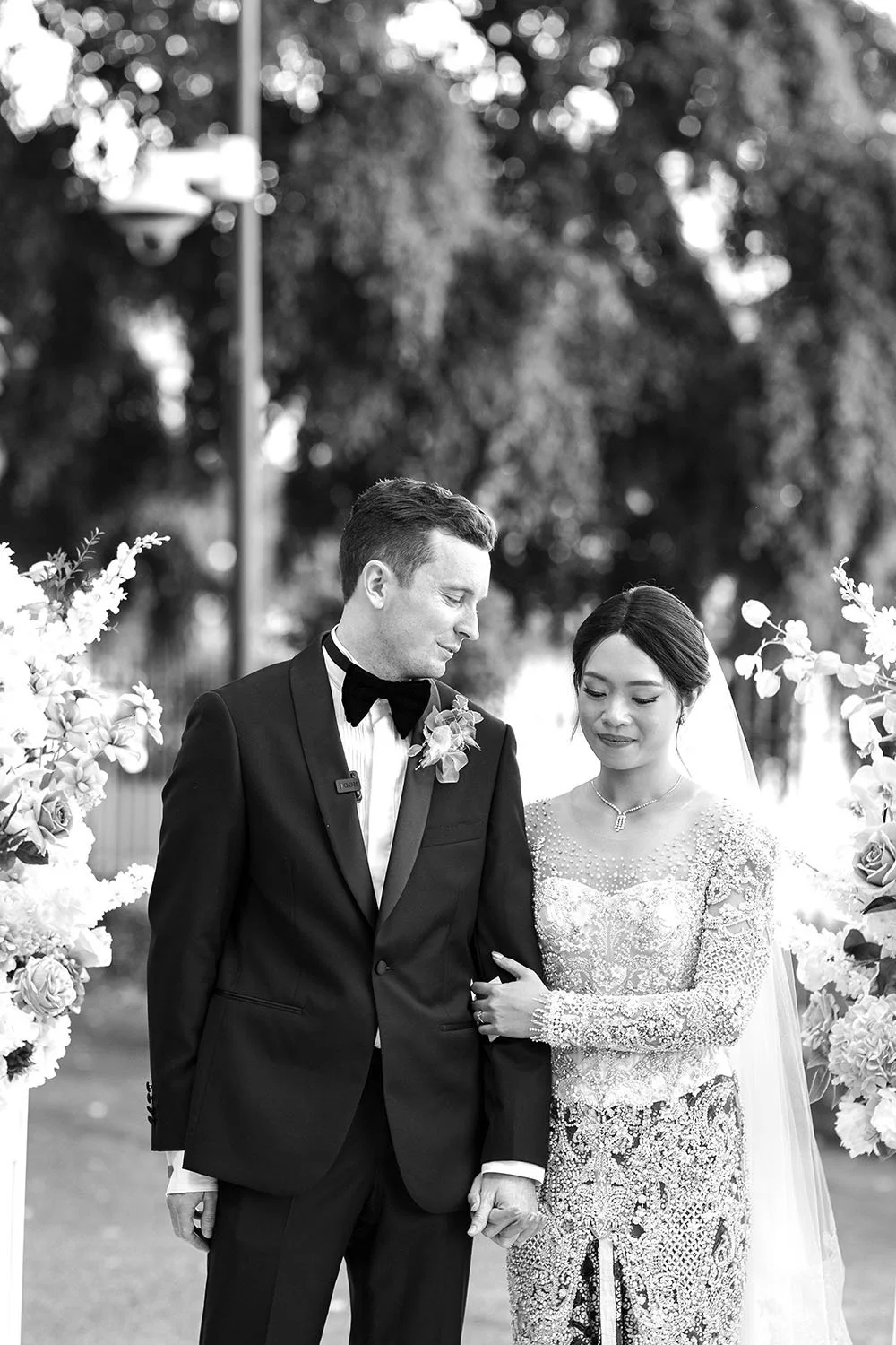 The bride Nori holds onto the groom Cliffe's arm and hand, looking down with a gentle smile