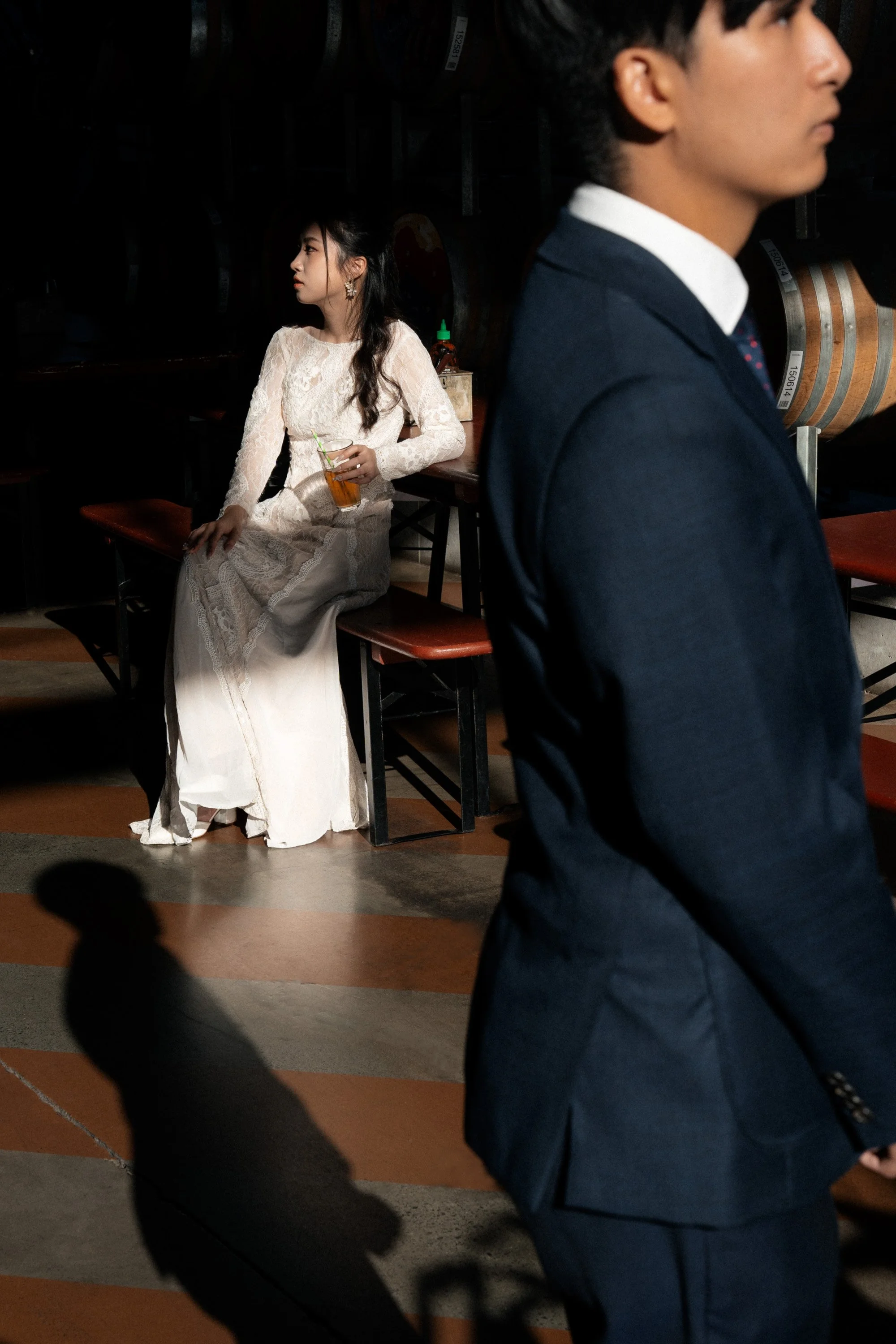 groom as a foreground look his right side with a shadow on the floor, bride Cassie as a background sitting on the bench holding a drink