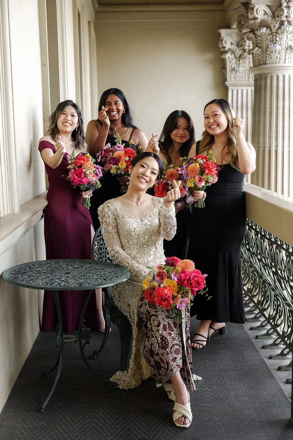 bride Nori sitting on the chair and her brides mates stand behind her and they both doing a love hand gesture to Whistler Li