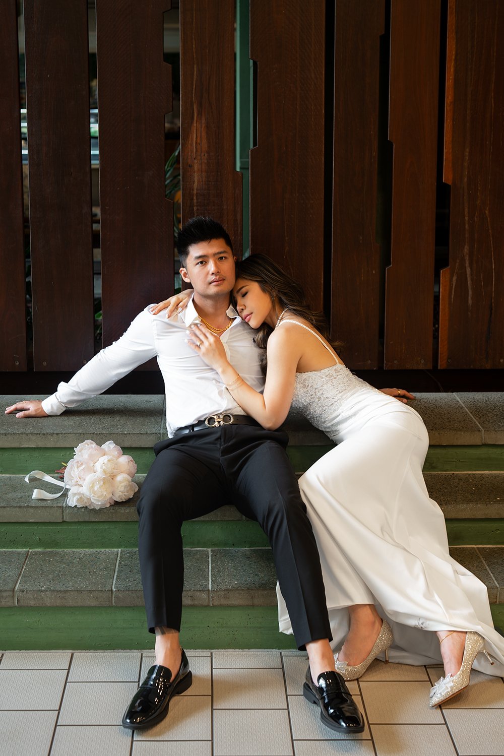 Groom Kai sitting on the stairs with both hands supporting him, while the bride Tiffy leans gently against him with her eyes closed, creating a tender and intimate moment. The bride wears a delicate wedding gown, and the groom is dressed in a classic