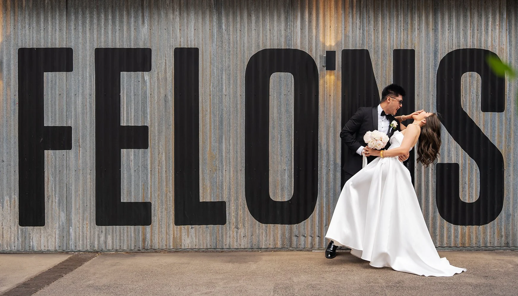 Groom Kai dipping the bride Tiffy as he leans in for a kiss, while the bride playfully covers her face with her hands and tilts her head back in front of the Felions Restaurant signage. The fun, romantic moment is captured with the restaurant's logo 