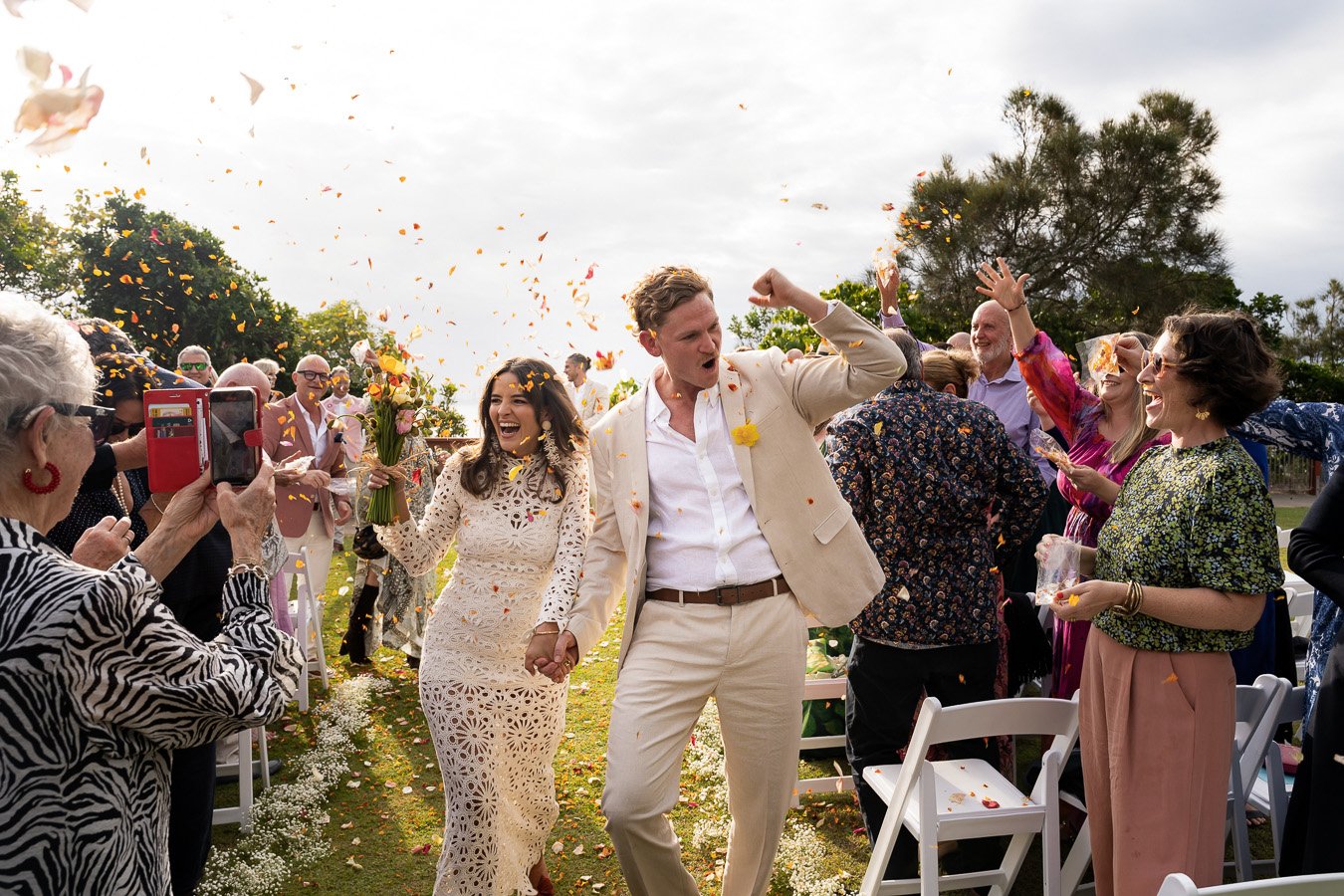 Lucy holding Tim's hand walking down the aisle, their family and friends throw paddles to the air cheers for them, everyone is happy captured by Brisbane wedding photographer Whistler Li