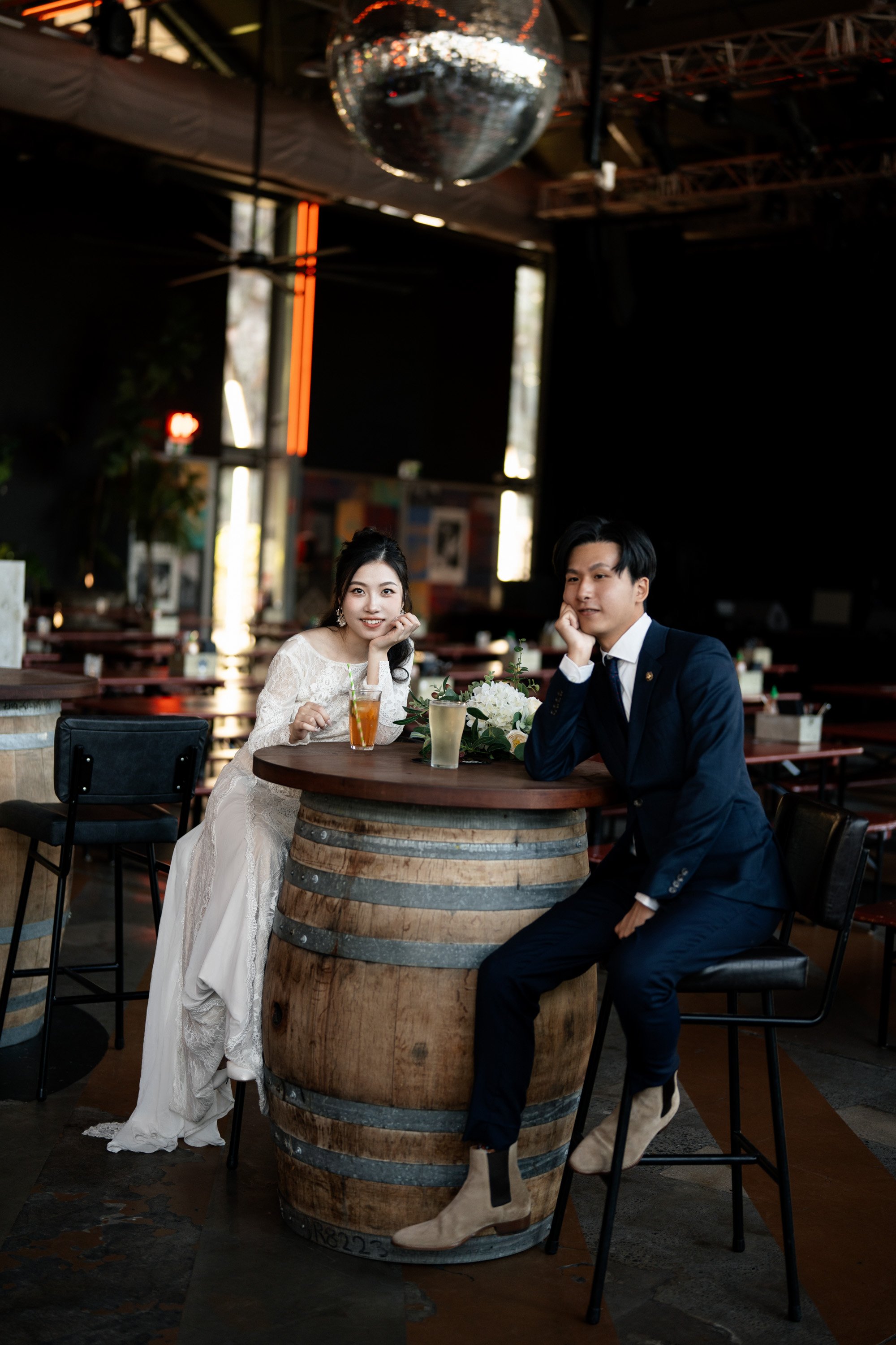 Bride and groom sitting next to a wine barrel, resting their hands on their heads as they look at the Brisbane wedding photographer. The relaxed pose and the rustic charm of the wine barrel create a casual yet intimate moment, capturing their connect