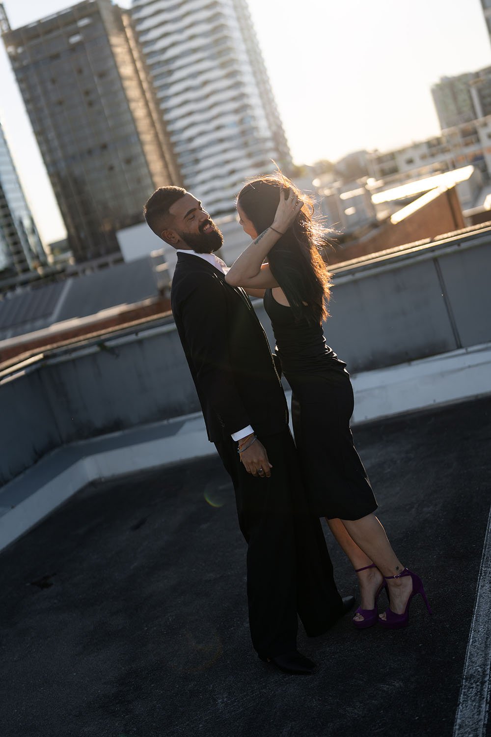 Jasmin pulls back her hair and smiles at Jay under golden hour light on a rooftop car park in Brisbane engagement photo session capture by Brisbane wedding photographer Whistler