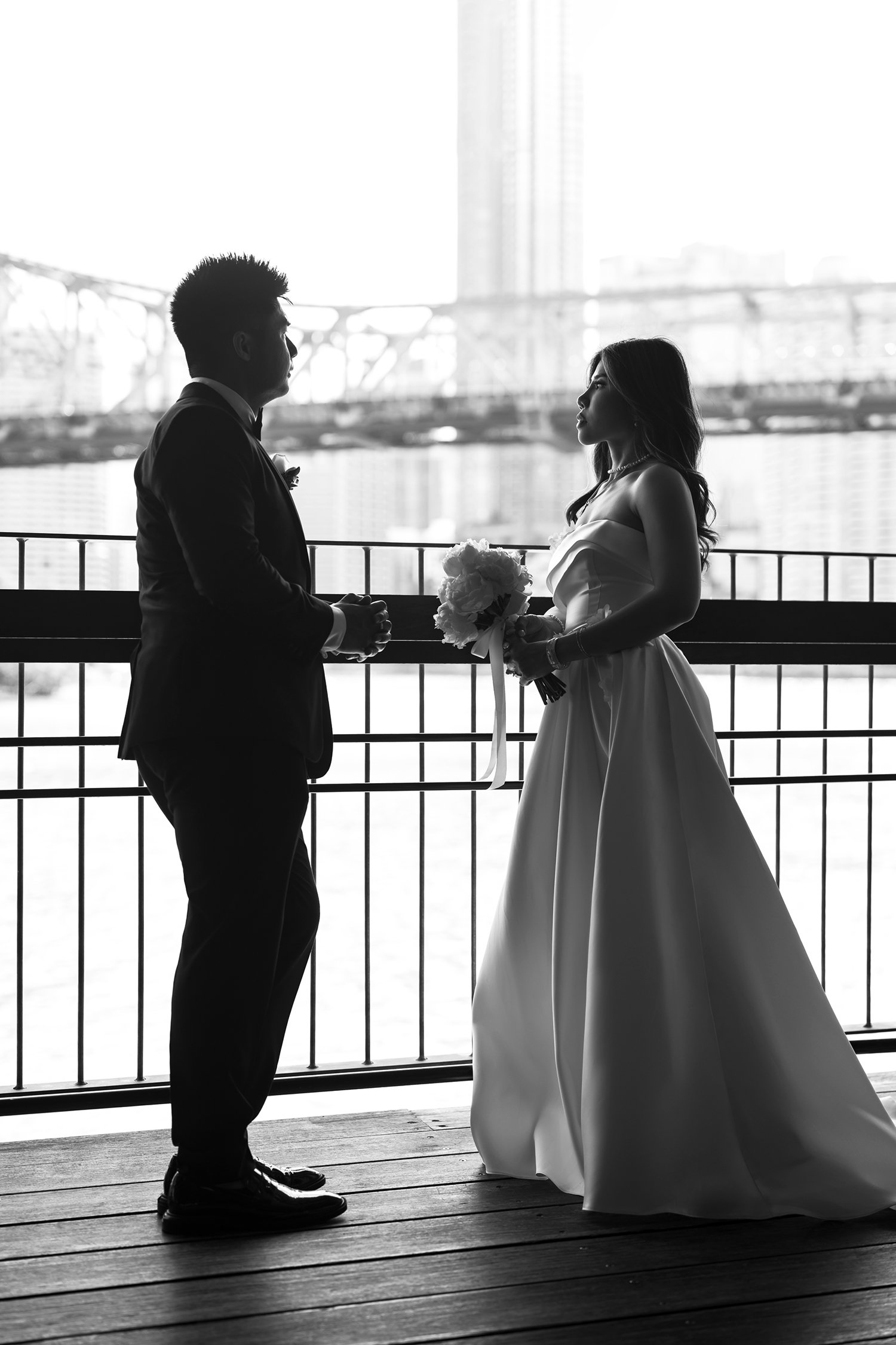 Bride Tiffy and groom Kai standing facing each other, gazing into each other's eyes, with the Brisbane River and Story Bridge in the background. The couple's connection is beautifully captured against the stunning cityscape, creating a romantic and i