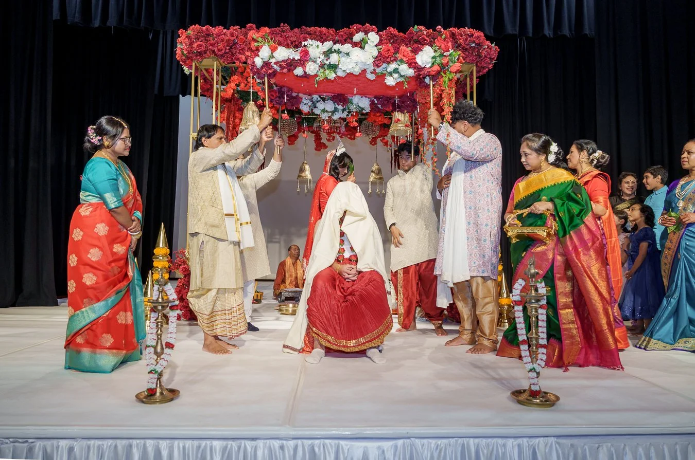 Amrita holding 3 leaf in front of her face walk around Casey, four males holding a floral Mandap