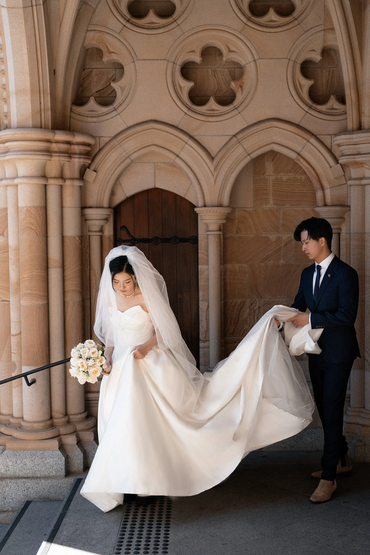 groom Leo holding Cassie's dress look at her outside St John's Cathedral in Brisbane