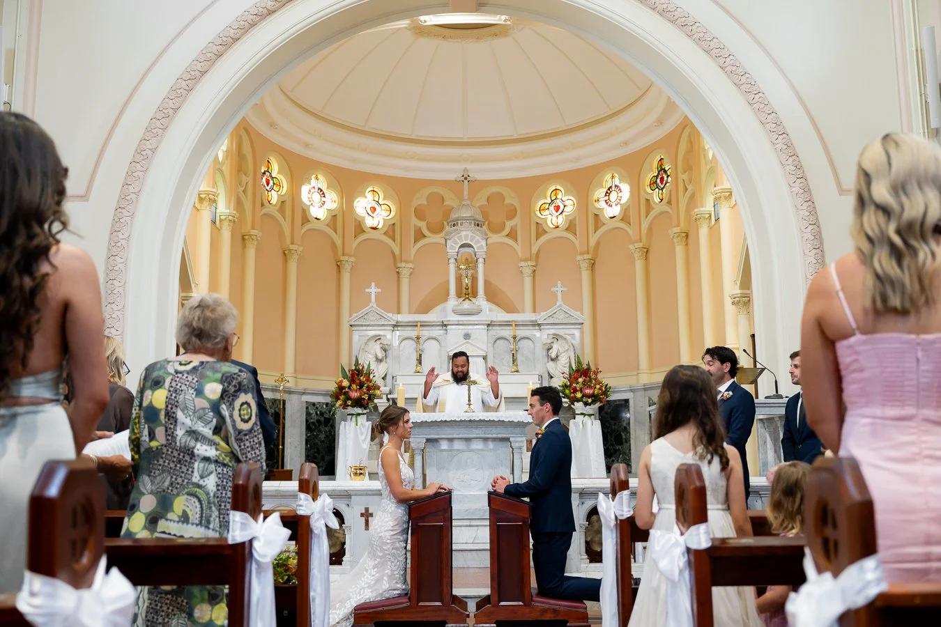 all guest standing, The bride and groom kneel before a small table, facing each other, while the priest stands at the altar in the distance.