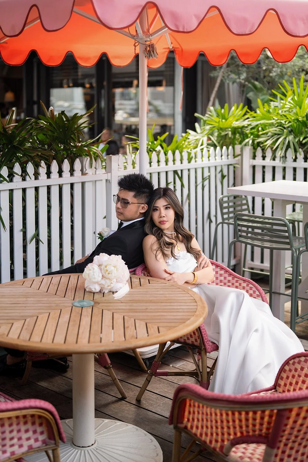 Tiffy and Kai seated together on a rustic wooden bench in a garden setting, surrounded by vibrant greenery. The bride wears a white lace wedding dress and smiles warmly, holding her bouquet, while the groom in a tailored black suit gently leans towar