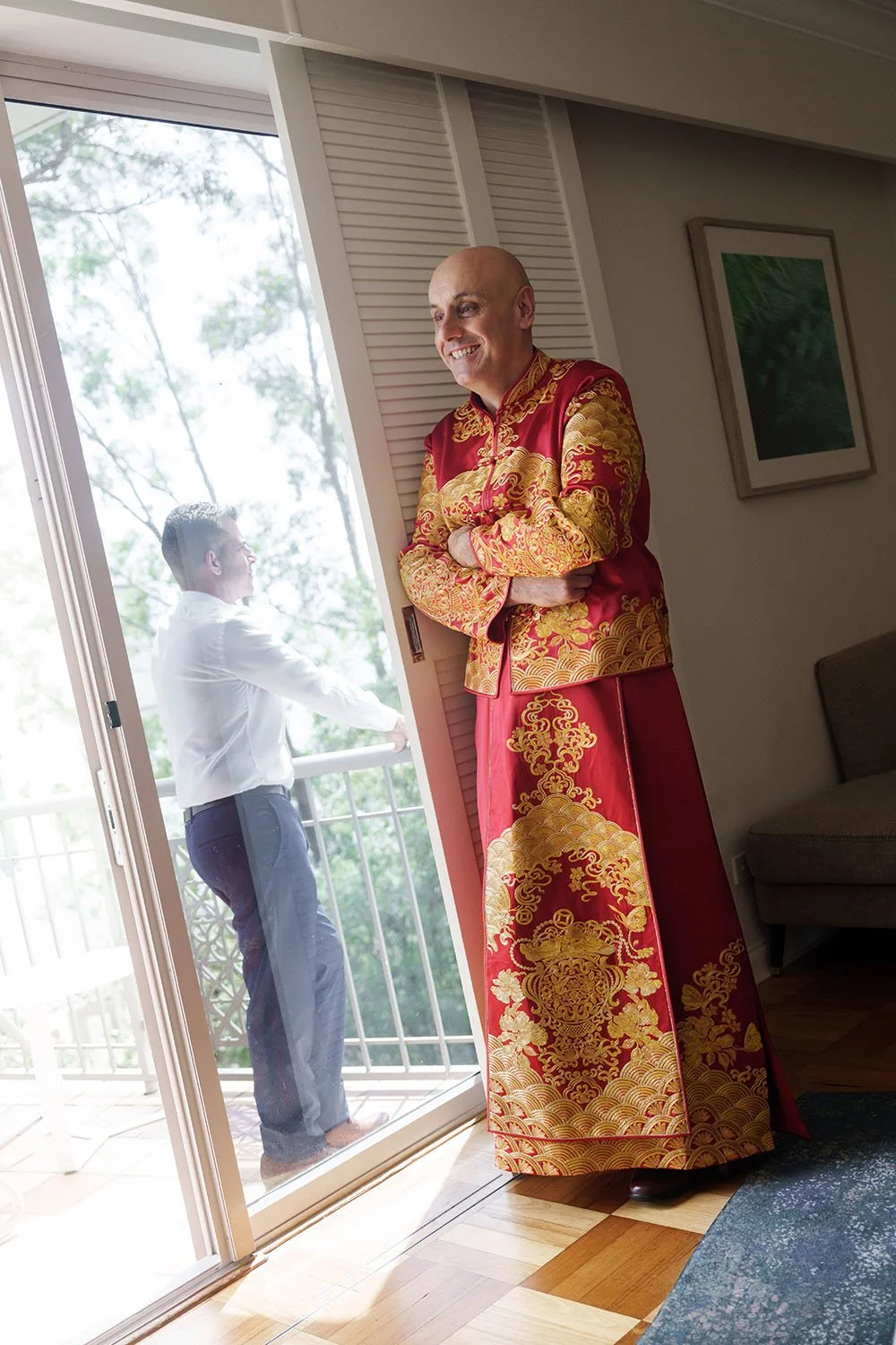 Robert stand next to the window crossing his arms in his traditional Chinese outfit and his groomsman stand on the balcony