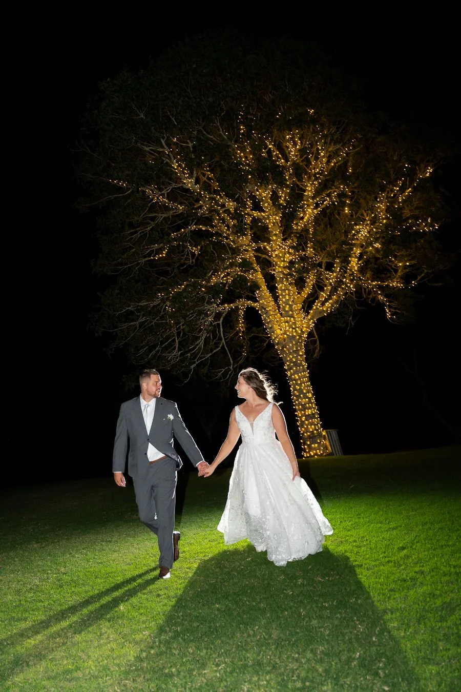 Chiarra and Ben holding hands walking on the grass at night, there is a fairy tree and flash light, lighting behind them