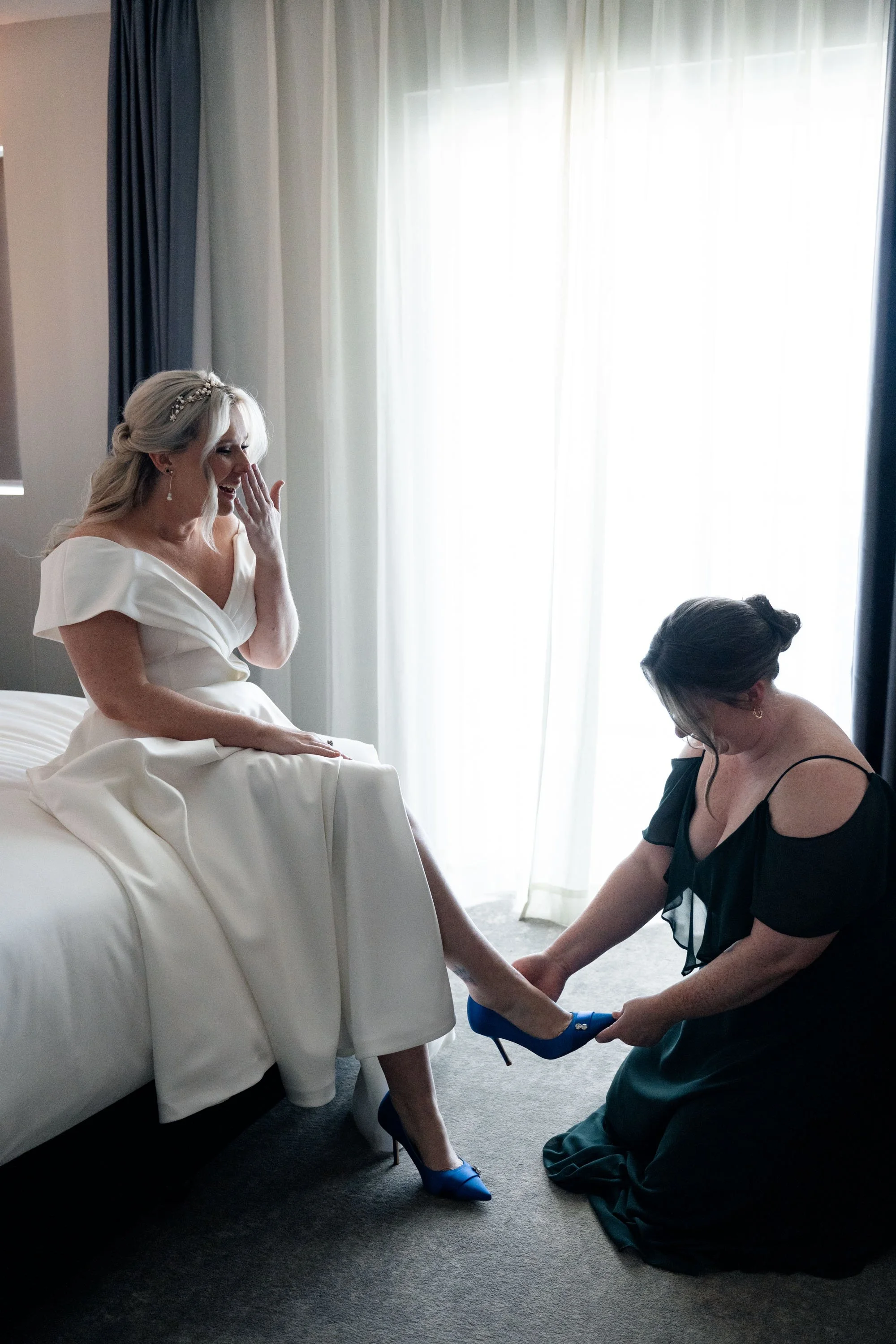 Bride's sister kneeling down to adjust the bride's high heels, offering a moment of care and support before the wedding ceremony