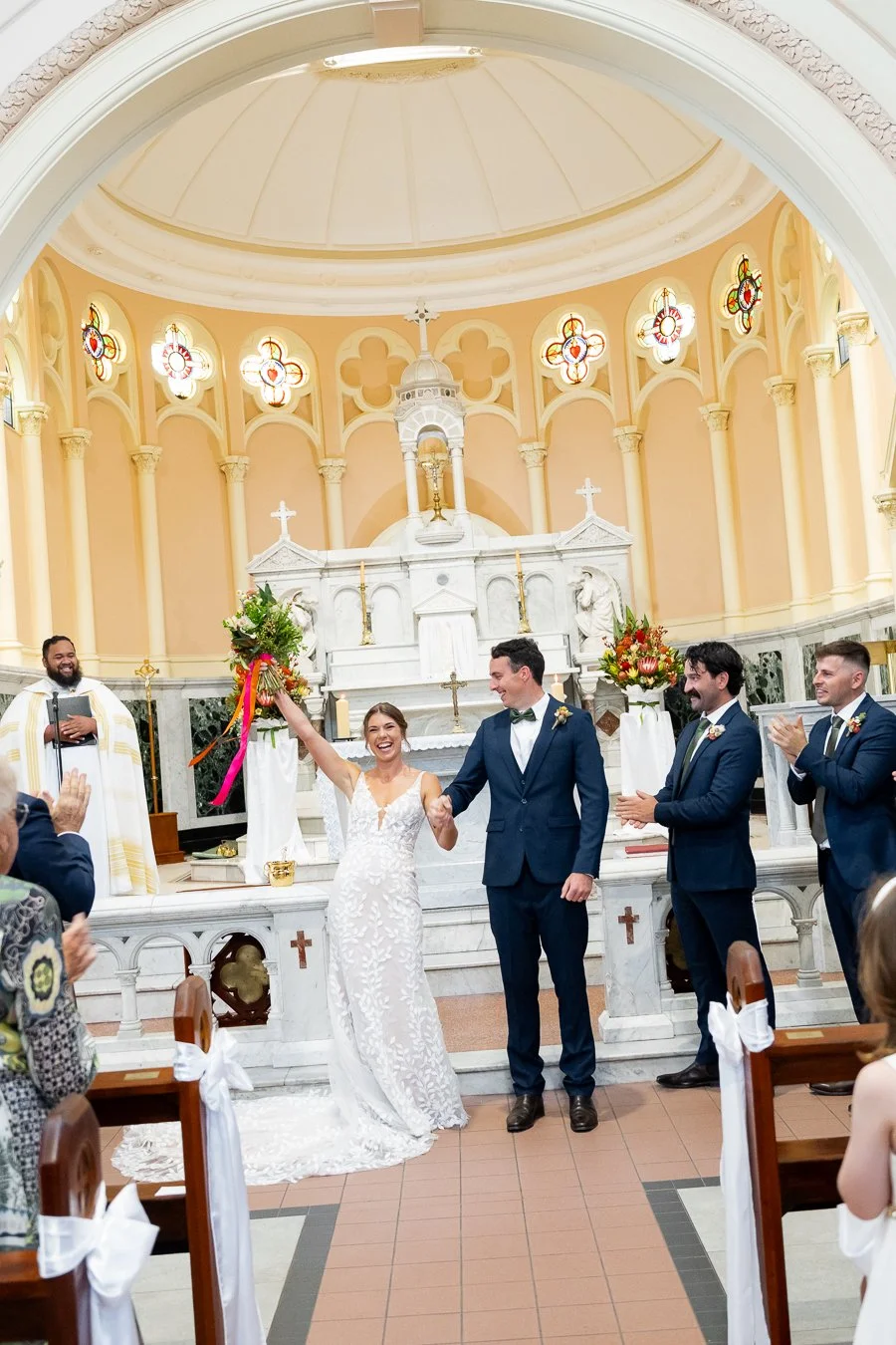 bride hands up with lot of smell while holding groom Giordan's hand, bridal party claps for them