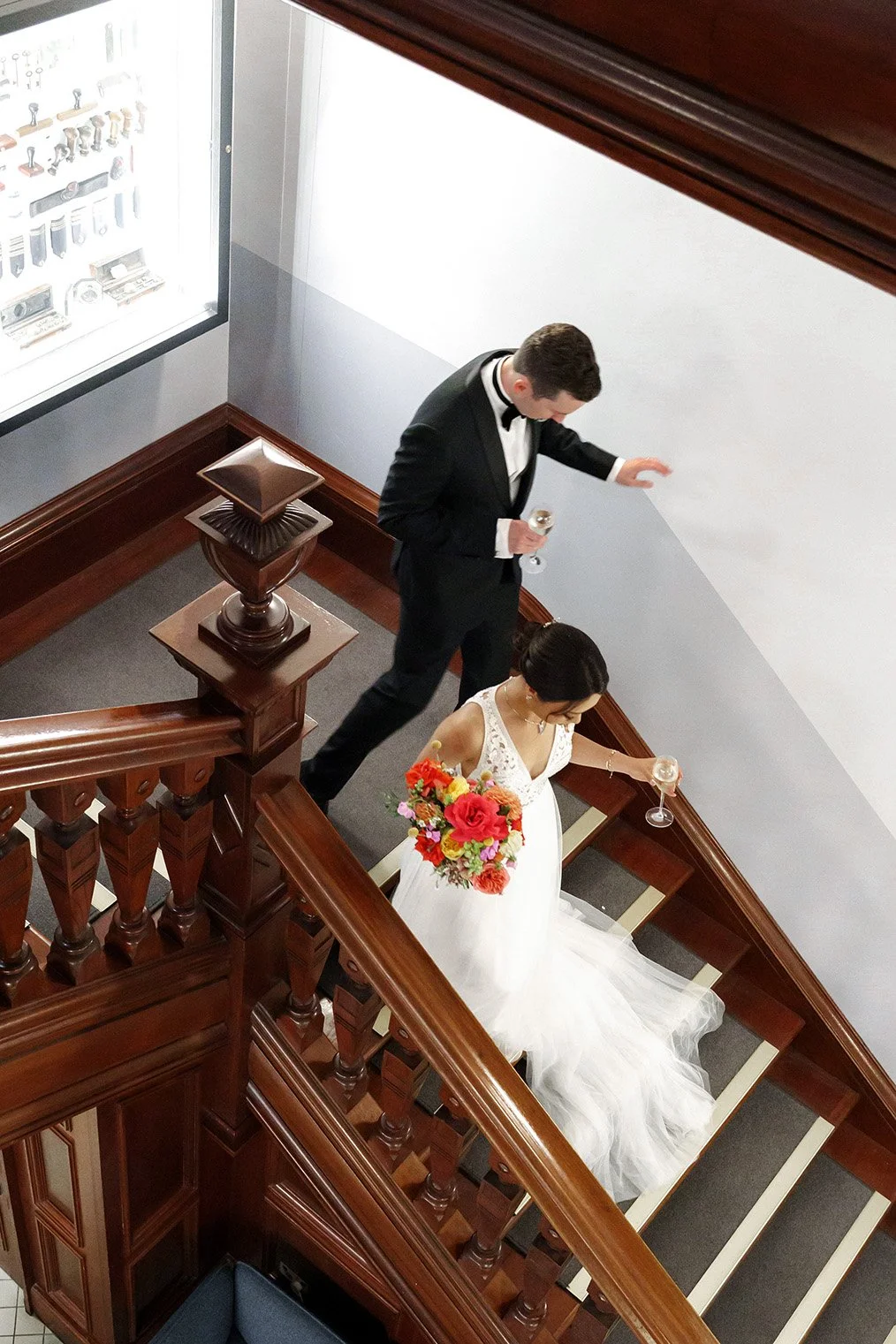 bride Nori holding a glass of champagne and her flower waling down the stairs in customs house Brisbane and groom Cliffe holding a glass of champagne as well walking behind her