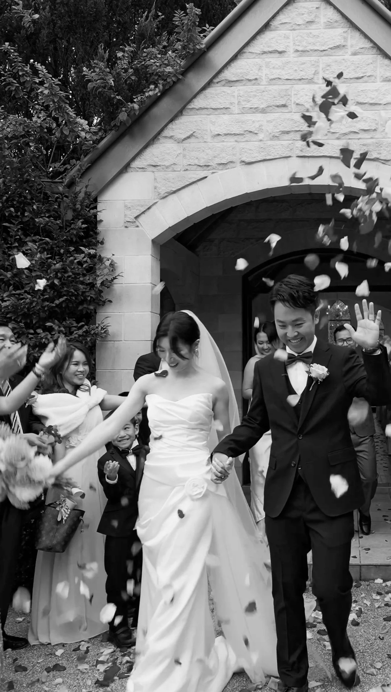 Flora and Jackey holding hands walking out of the chapel, guests in two lines throw rose paddles up to their top