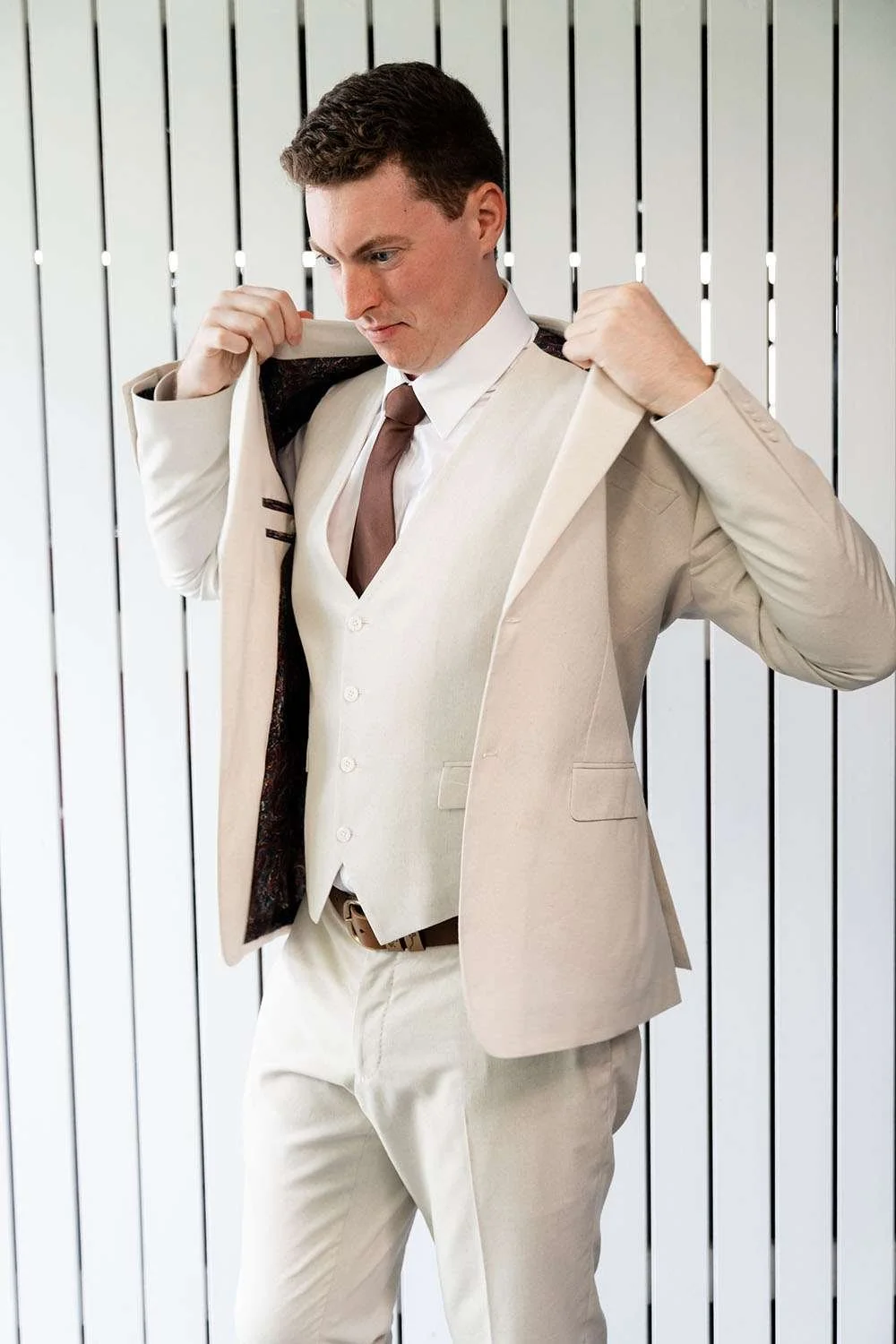 groom Jack put on his jacket by himself in front of white wood panel wall