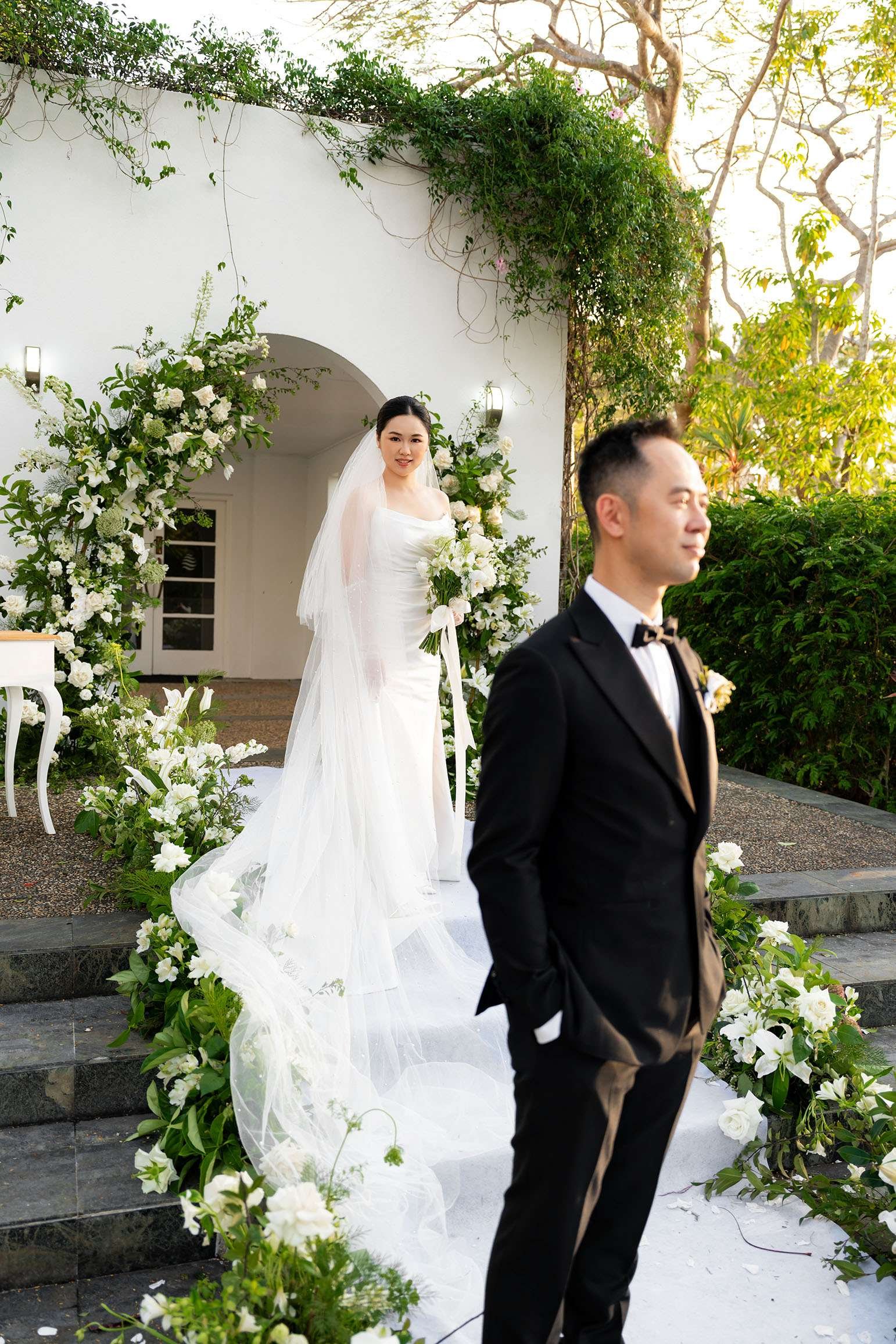 bride Wendy look at the camera, left holding a flower, groom stand few steps in front of her