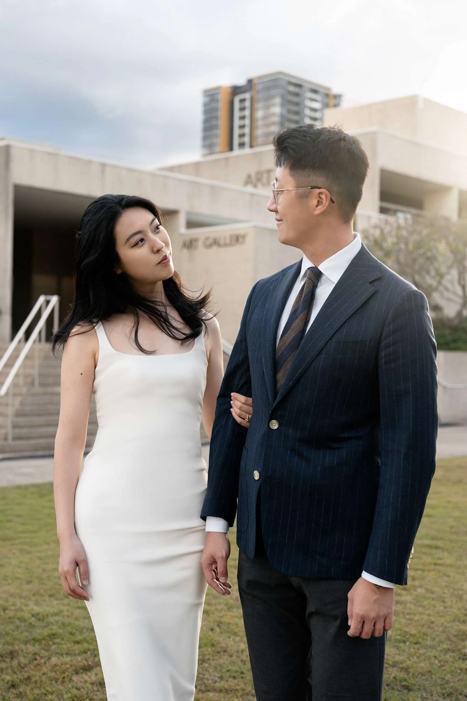 Kath holding Dave's arm as they look at each other, sharing a tender and intimate moment in front of an art gallery, with the architecture adding a sophisticated backdrop