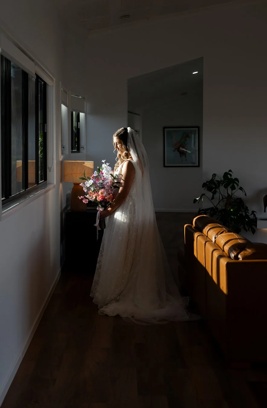bride Chiarra stand in-front of window look down to her flower and light hits her face