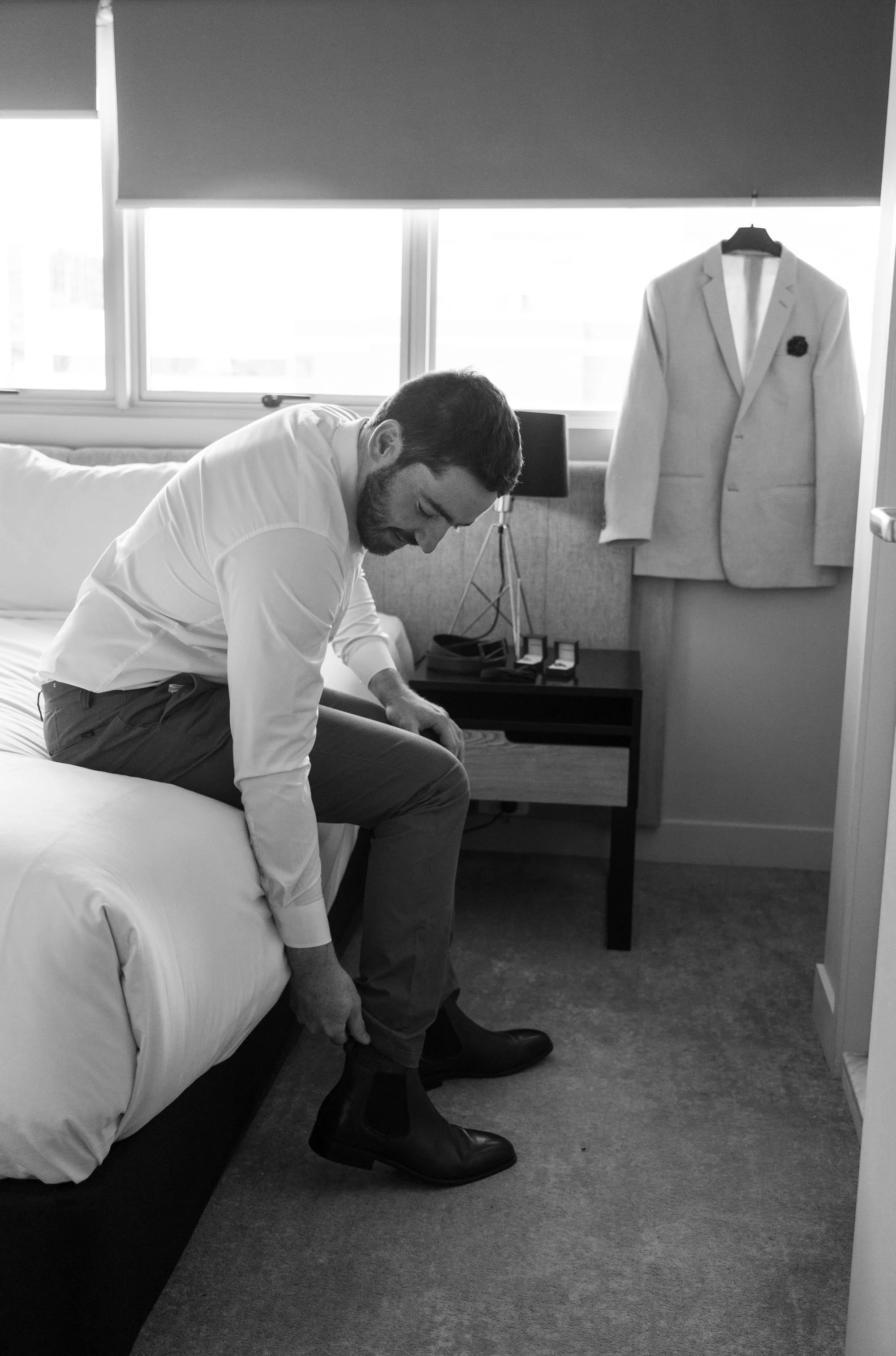 Groom Thomas adjusting his boots while sitting on the edge of the bed, preparing for the wedding day with a calm and focused moment