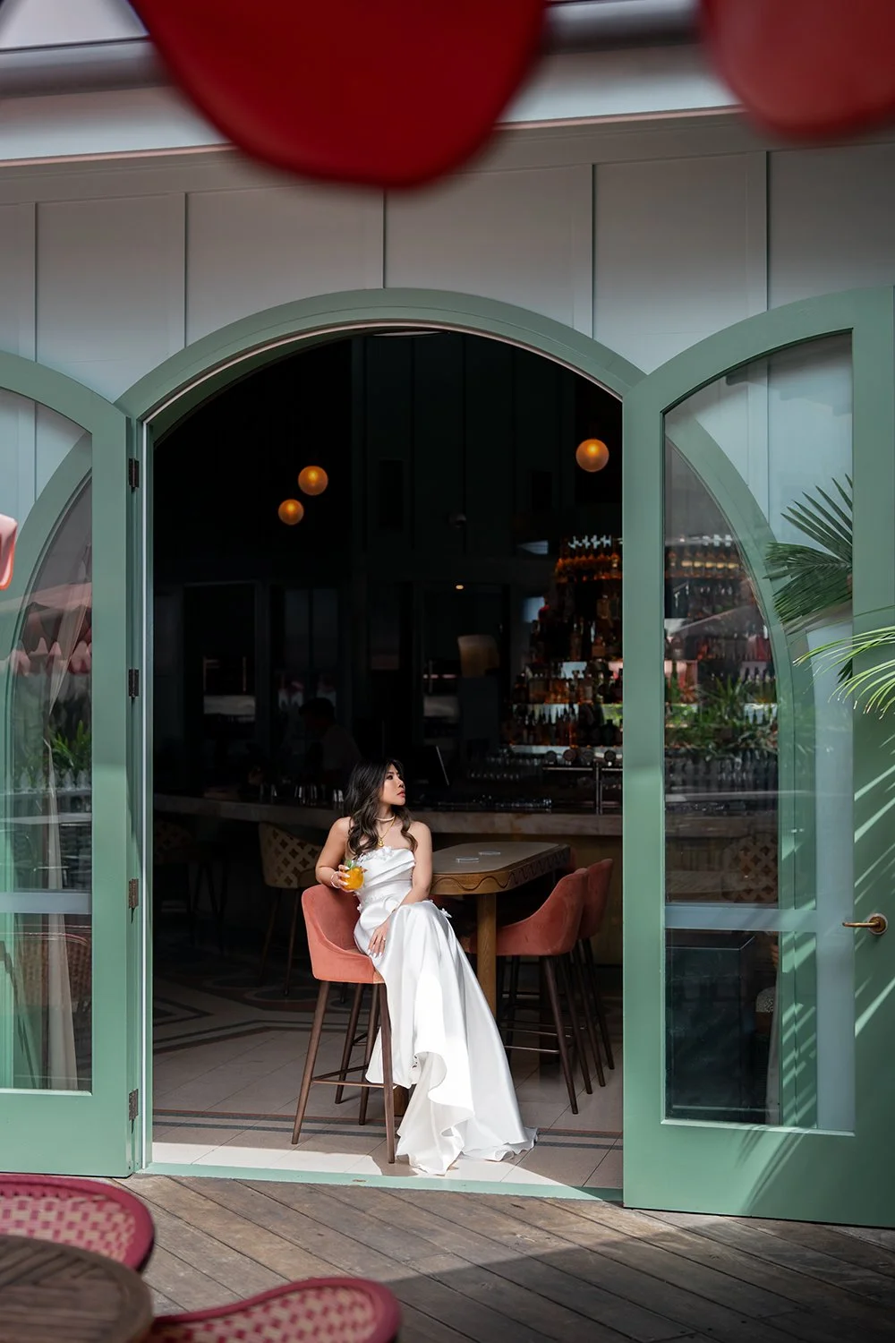 Tiffy sitting on a pink stool in a restaurant with green outdoor walls, holding a glass of fresh juice and gazing thoughtfully into the distance. She is dressed in a white wedding gown, creating a unique blend of elegance and casual charm in this mod