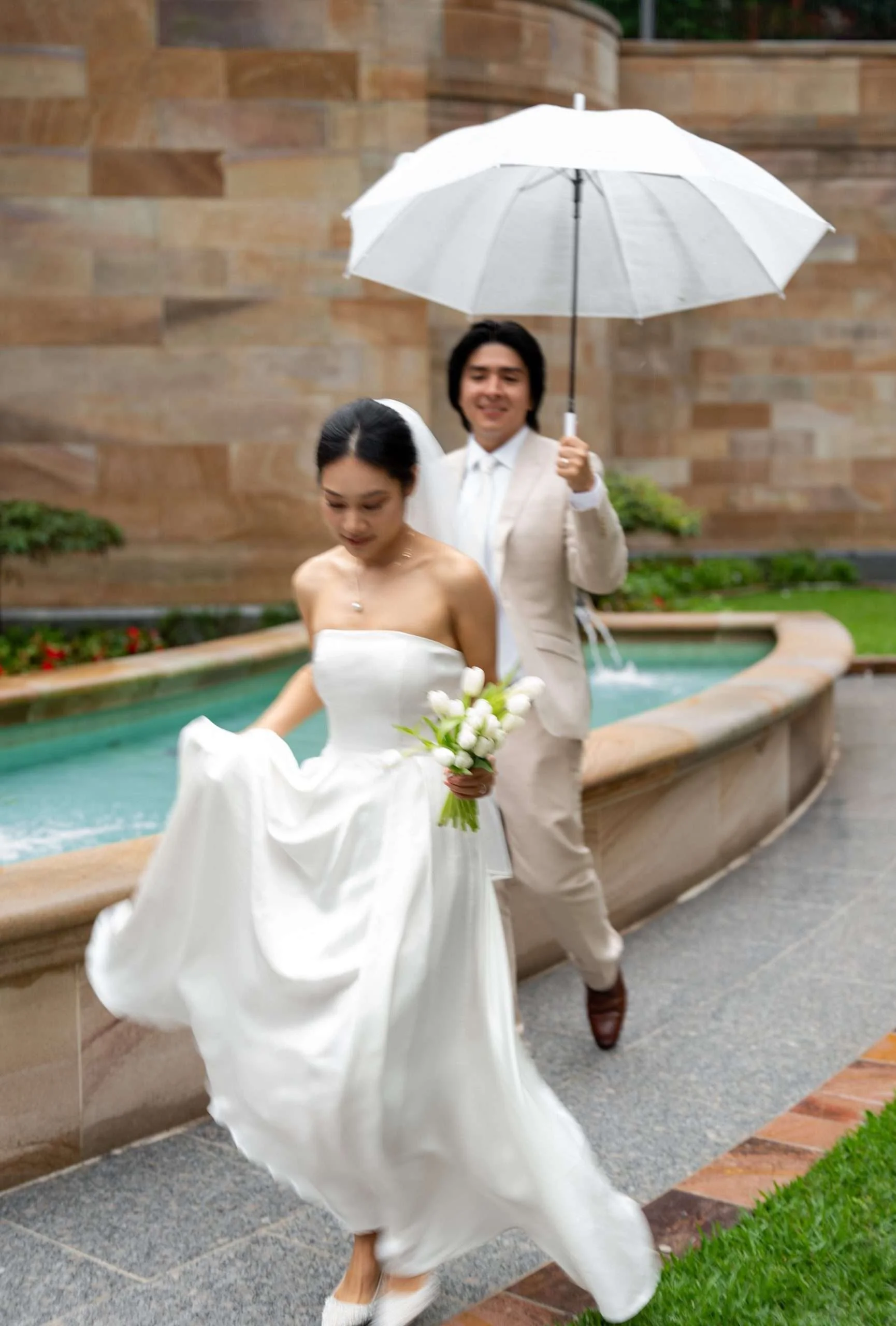 Yi holding her white dress and tulip running in the rain, and Jon holding a white umbrella running behind her