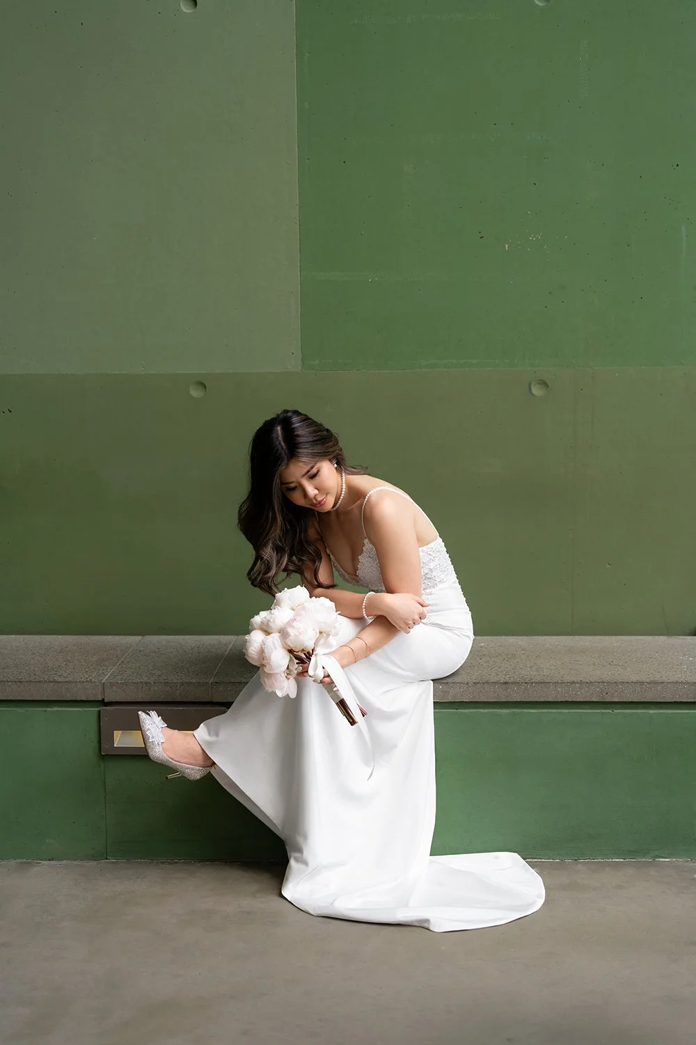 Bride Tiffy sitting down in front of a green wall, gazing down at her bouquet of flowers. She is wearing a beautiful wedding gown, with soft natural light highlighting the delicate details of the flowers and her serene expression, creating a peaceful