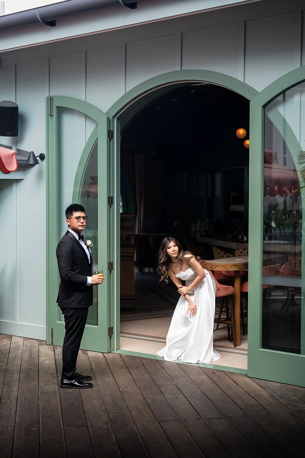 Tiffy and Kai standing outdoors on a sunny day, surrounded by lush greenery. The bride wears a flowing white gown with intricate lace details, holding a pastel bouquet, while the groom stands beside her in a sleek black suit, looking lovingly at her.