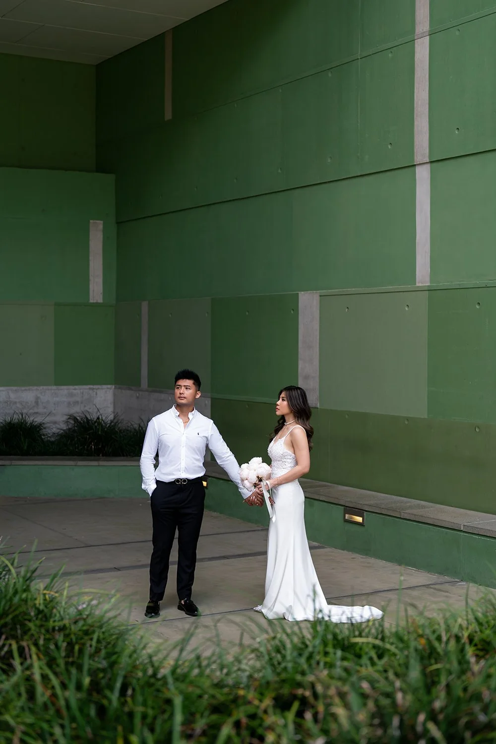 Groom Kai holding the bride TIffy’s hand as they both look away, standing in front of a vibrant green building. The scene captures a modern and stylish wedding moment, with the couple’s connection highlighted against the bold and colorful backdrop