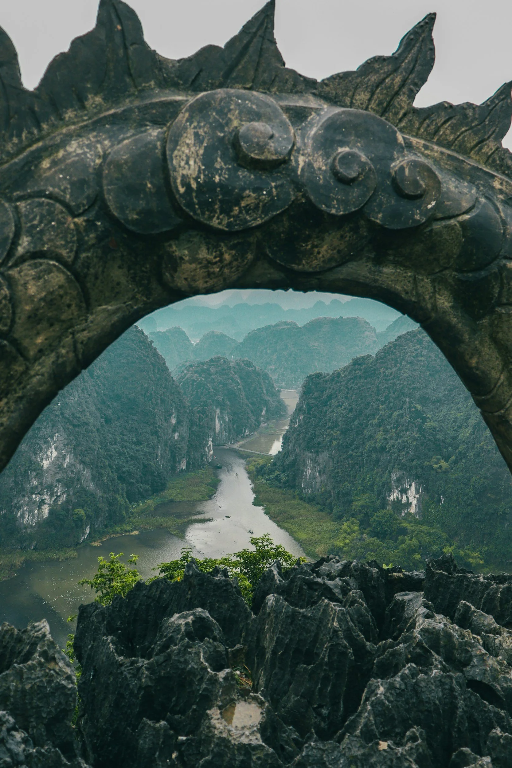 View of a river meandering through lush green mountains, framed by a carved stone archway with decorative spirals and leaf motifs at the top.