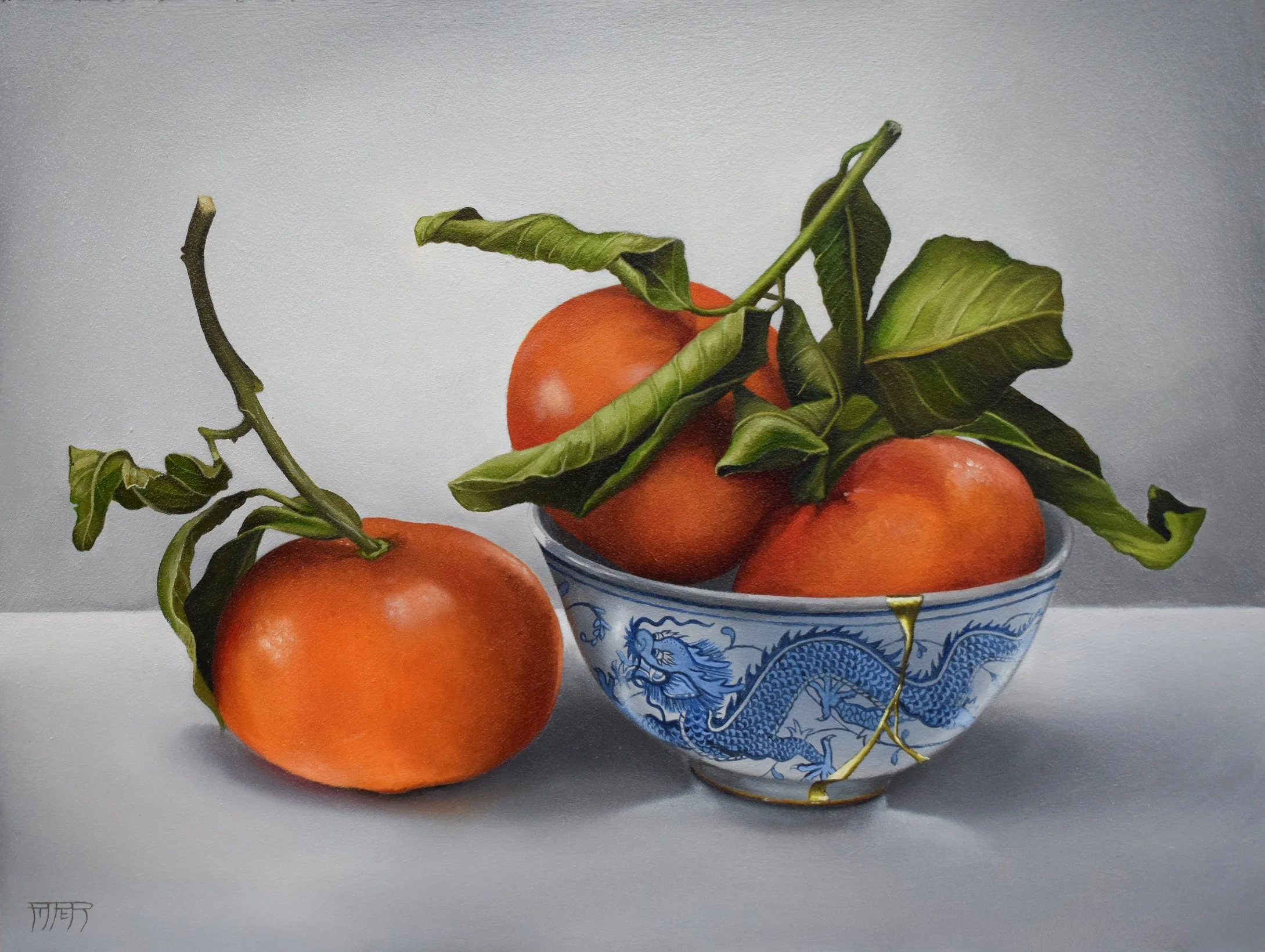A porcelain bowl with blue dragon patterns holds ripe oranges with green leaves and stems, placed on a white surface against a plain, light-colored background.