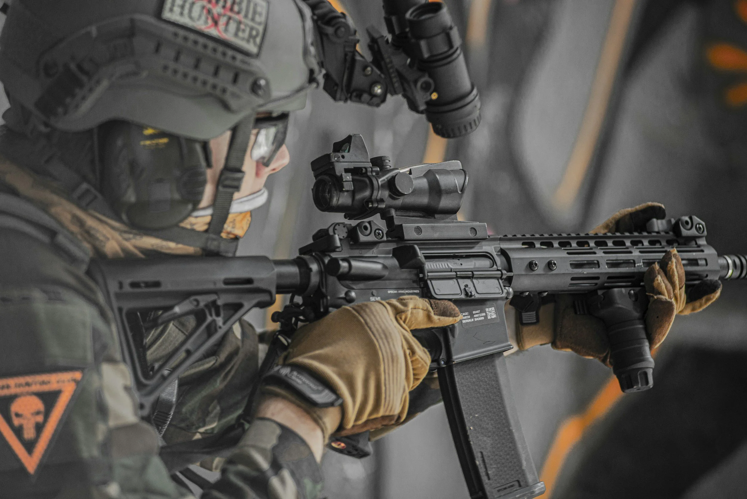En person i militäruniform och skyddsutrustning som tittar genom en siktesmontage på en vapen, inomhusmiljö.