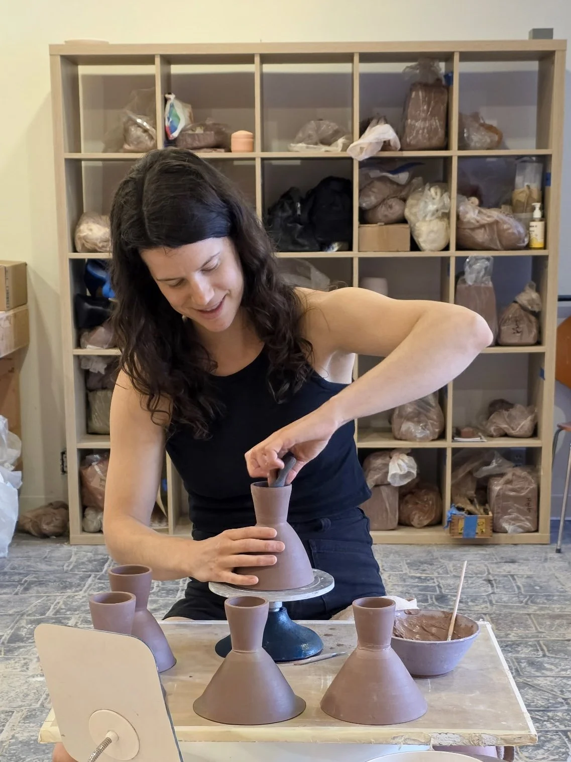 Woman shaping a clay vase on a pottery wheel, surrounded by finished pottery pieces, in a studio with shelves filled with clay and supplies.