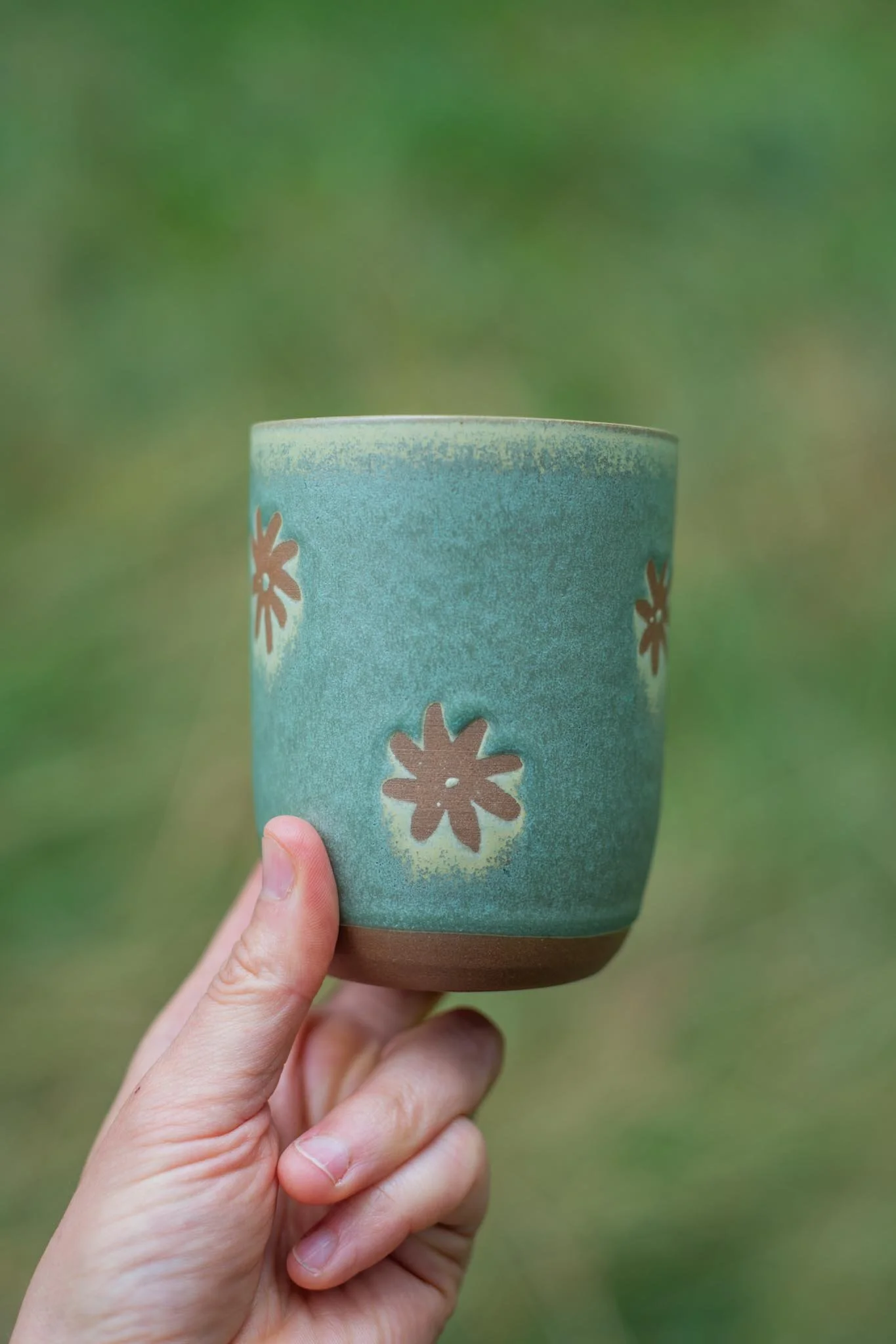 A person holding a ceramic cup with a teal exterior decorated with brown and pink flower patterns, with a blurred green background.