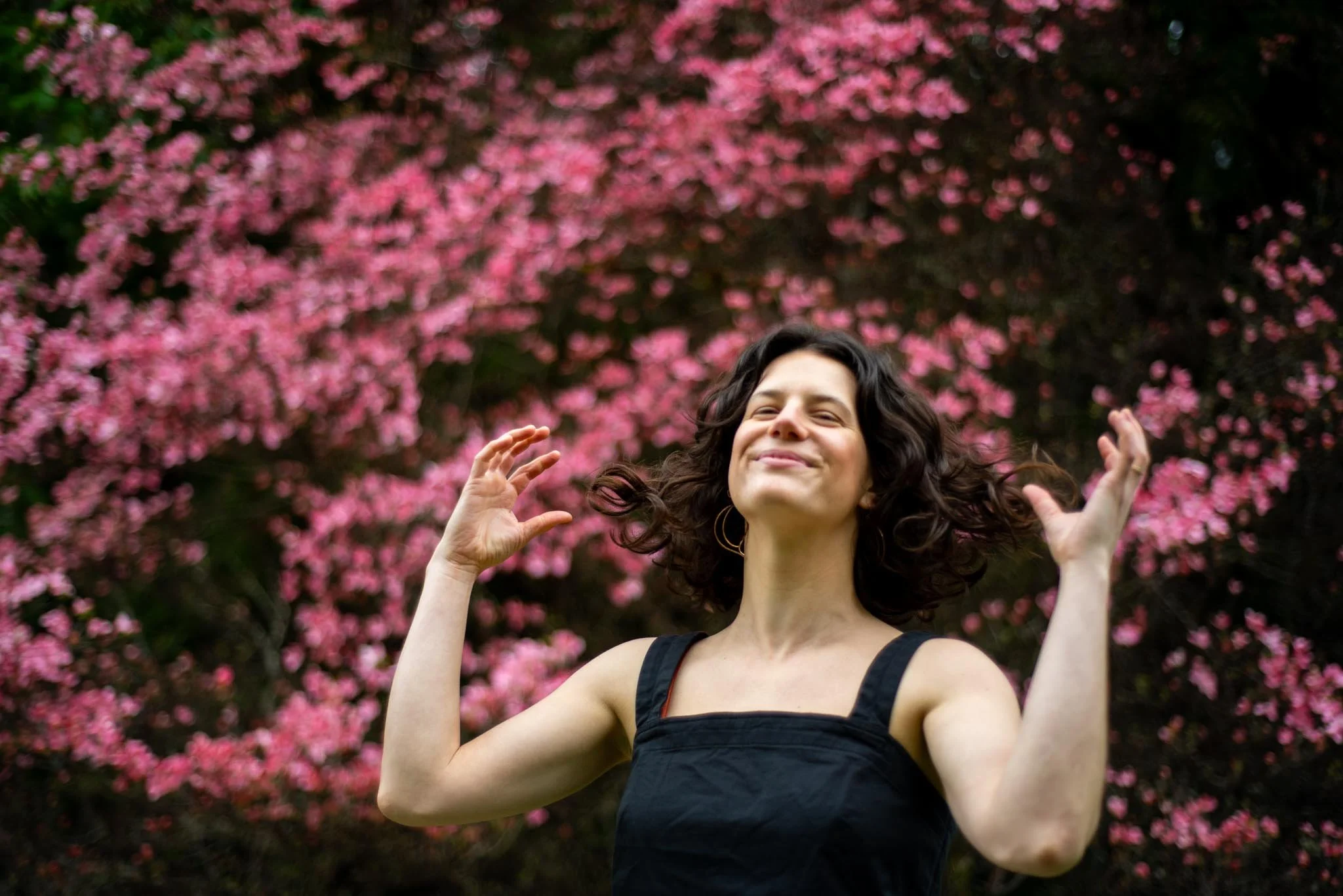 A woman with dark, curly hair smiling with eyes closed in front of a pink flowering tree.