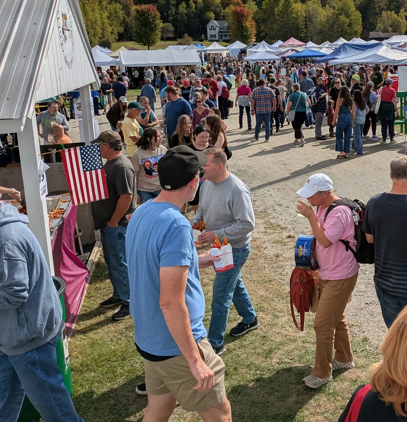 People walking outdoors at a fair with tents and booths, colorful trees in the background, and clear weather.