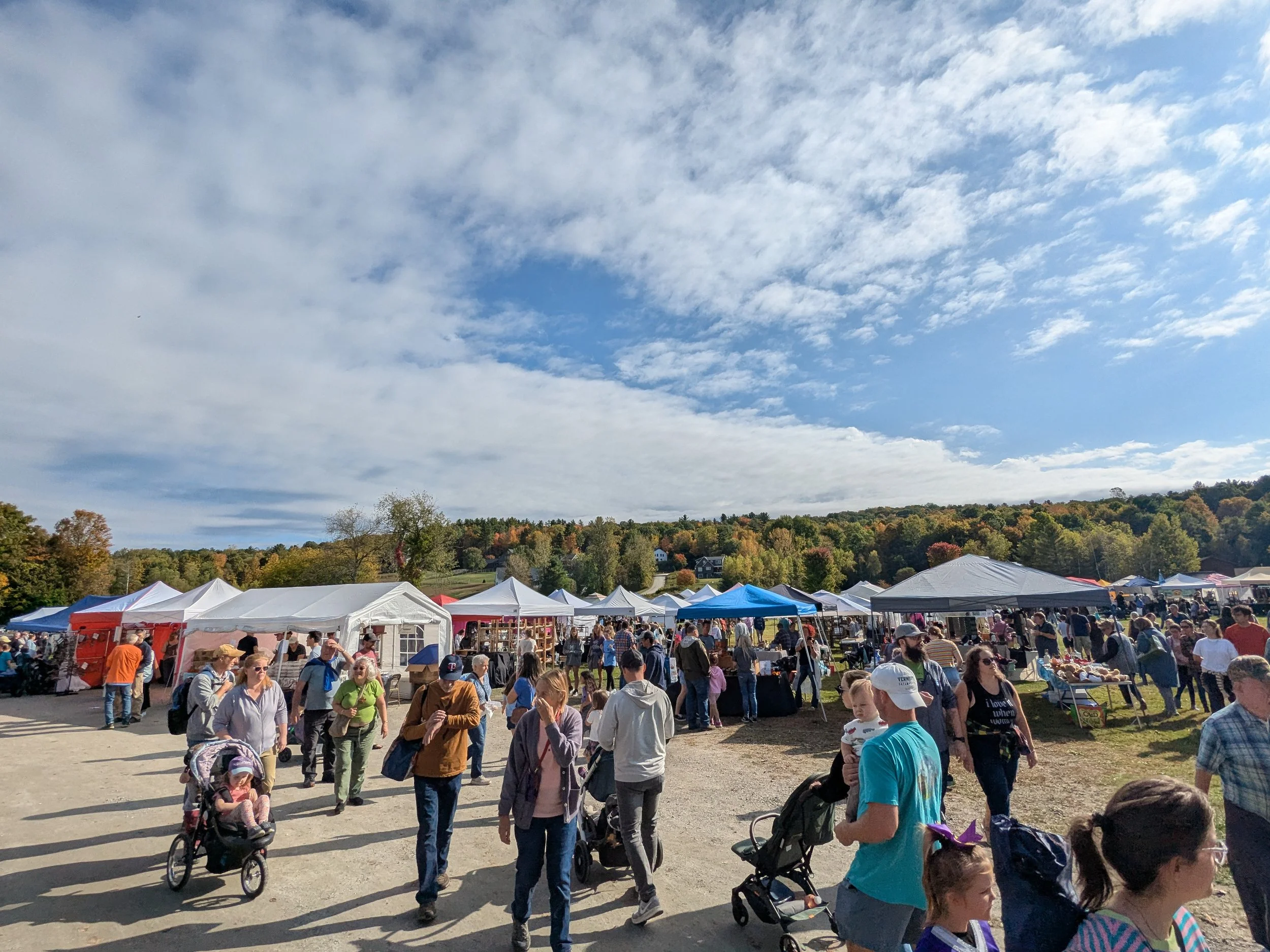 A busy outdoor flear market on a bright day with partly cloudy skies, bordered by trees with autumn foliage.